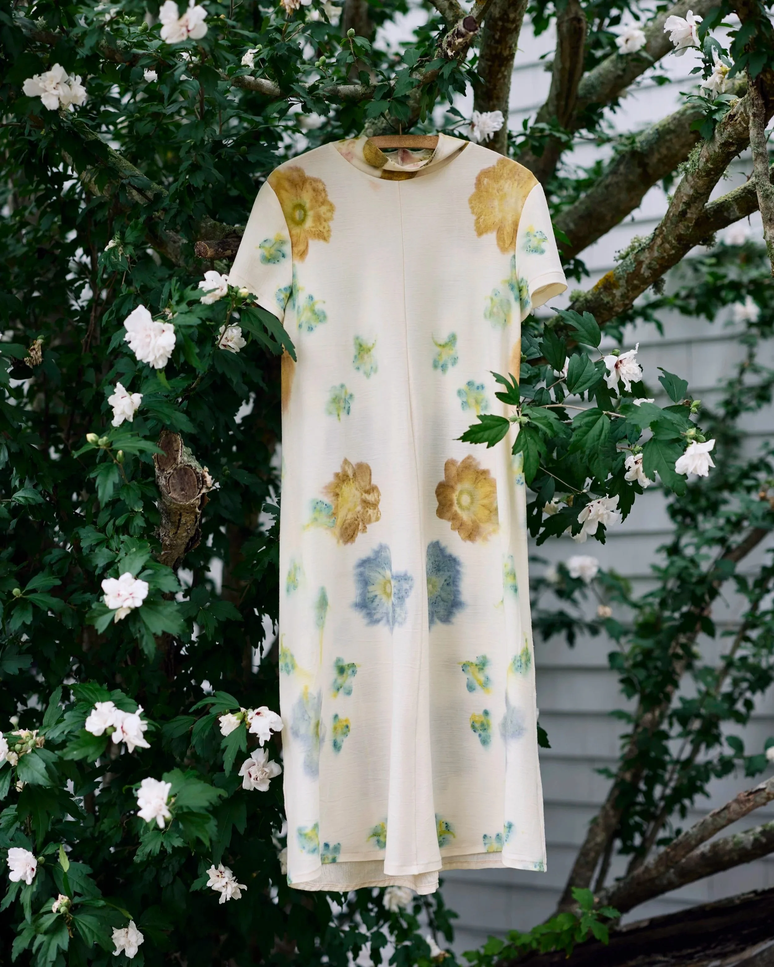 A cream-colored dress with large floral anemone eco prints patterns hanging from a hanger on a tree branch in a garden with white flowers and green leaves.