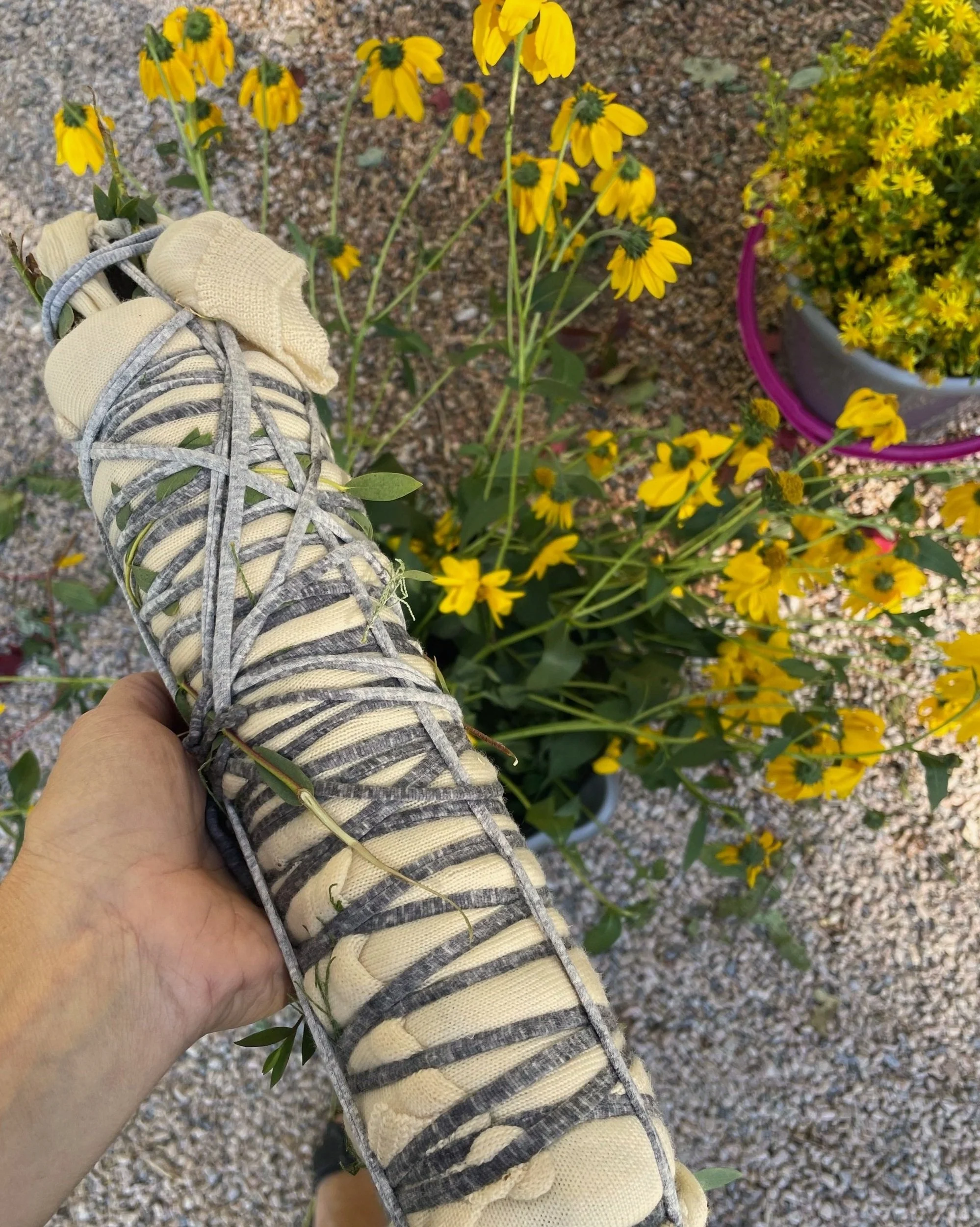 Person holding a hiking shoe with gray laces and cream-colored fabric. In the background, yellow flowers and a potted plant with small yellow blossoms.