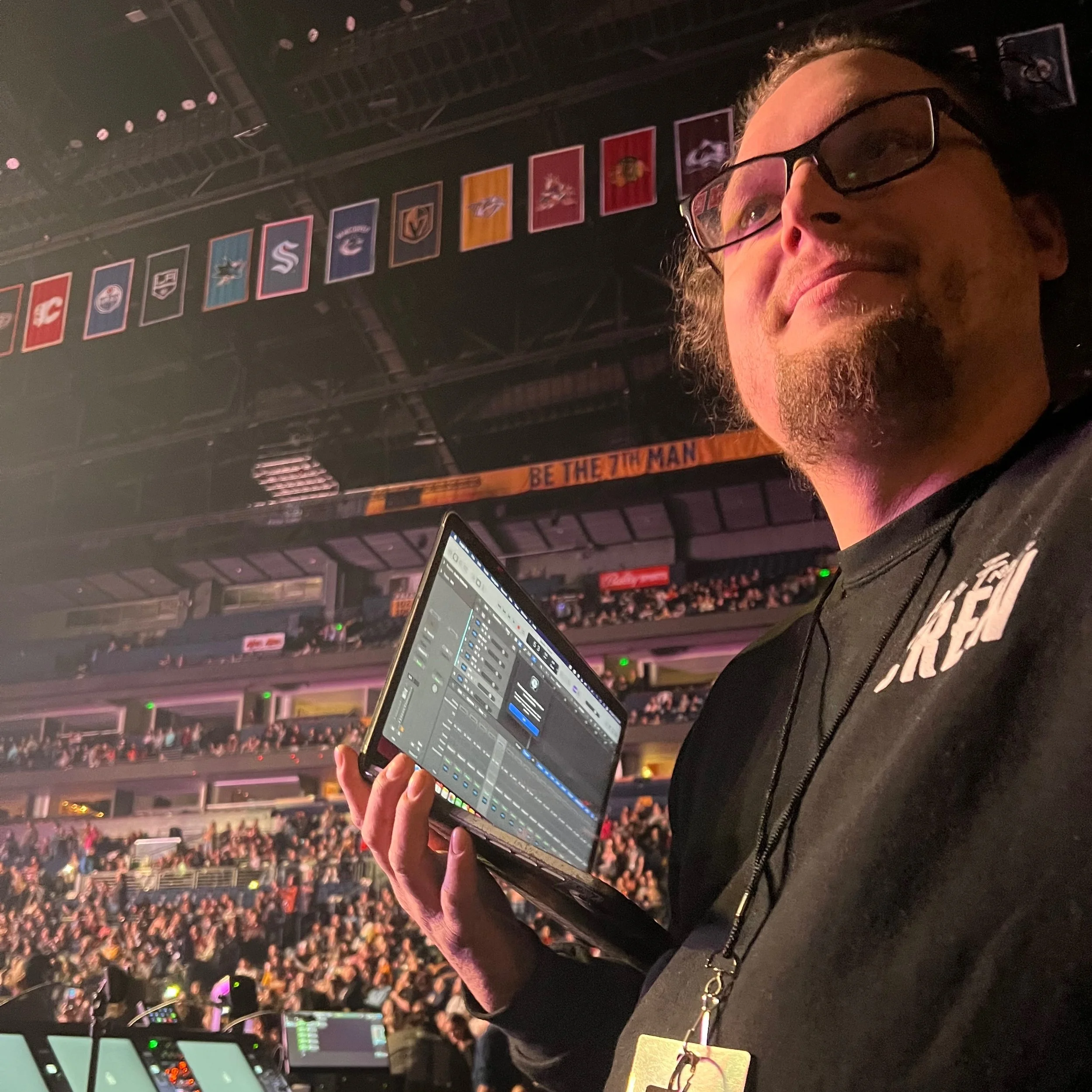 A man with glasses and a goatee holding a laptop at a crowded indoor event, with banners hanging from the ceiling and a sign that says "BE THE 7TH MAN" in the background.