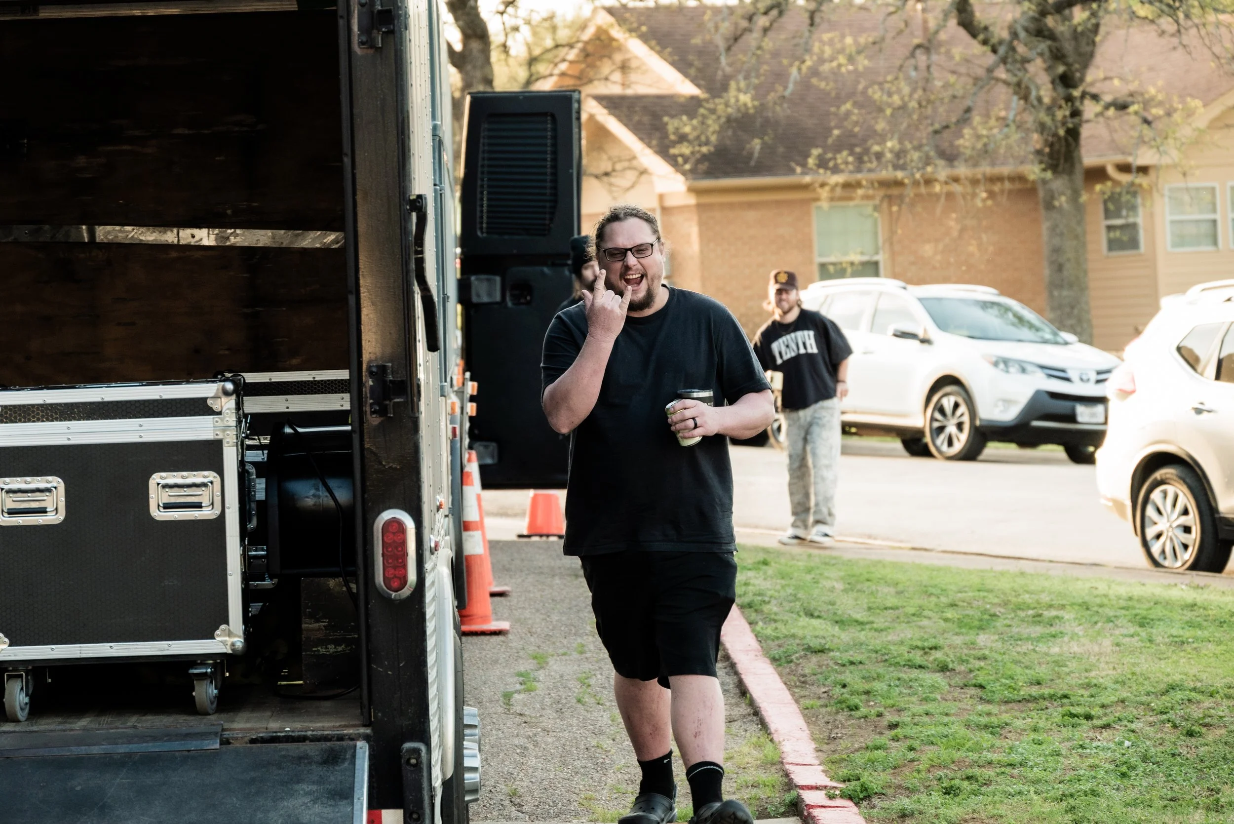 A man with glasses and a beard in a black t-shirt and shorts making a rock-and-roll gesture with his hand and smiling, standing next to a truck outdoors in a residential area with other cars and a house in the background.