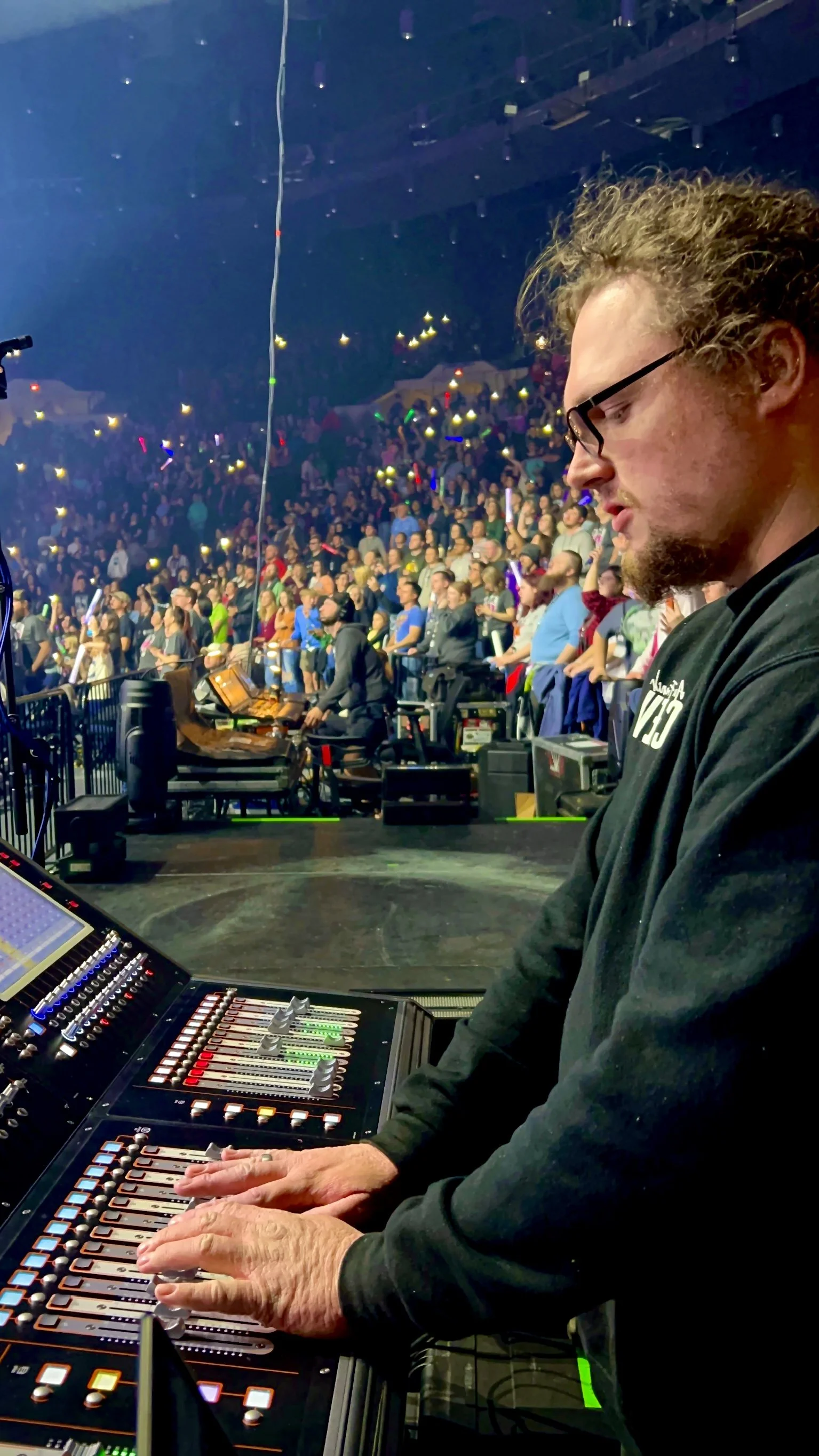 Sound engineer adjusting controls on a mixing console during a concert with a large audience in the background.