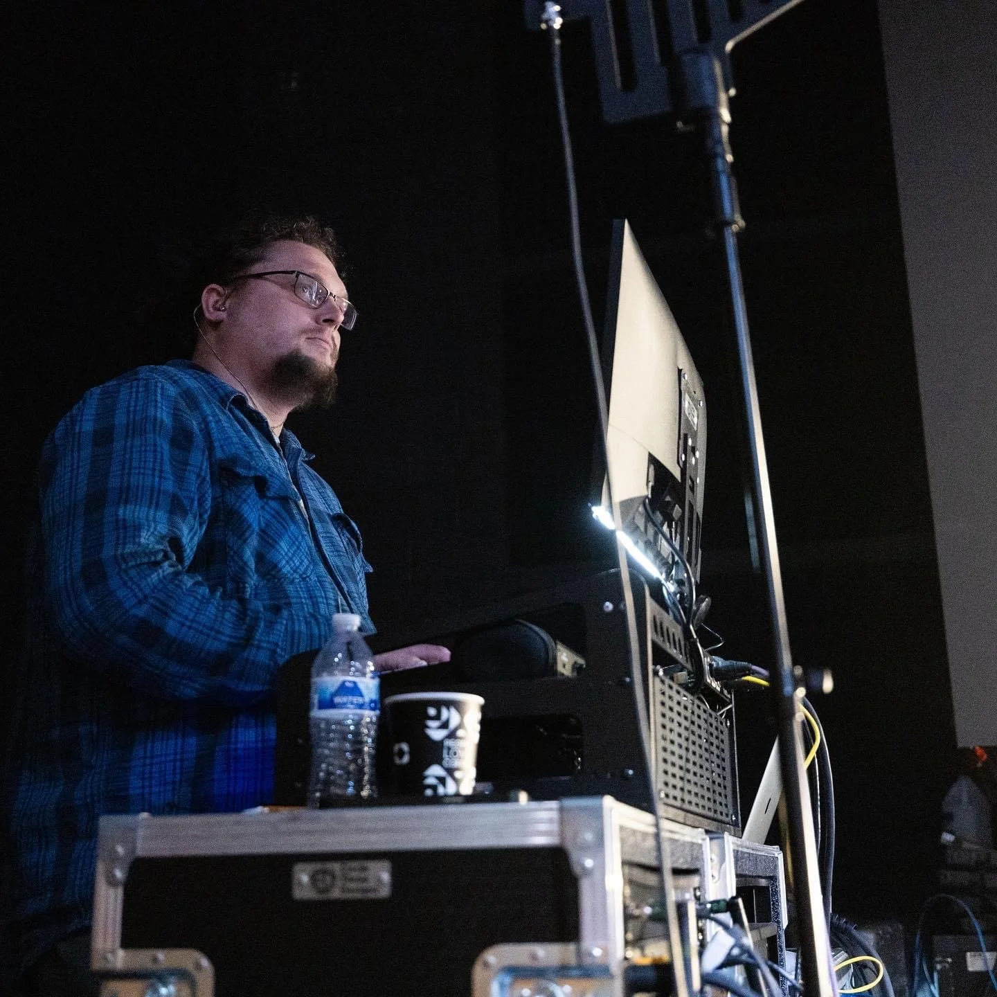 A man with glasses and a beard wearing a blue plaid shirt working at a computer station on stage, with a water bottle, a coffee cup, and various electronic equipment on a table in front of him.