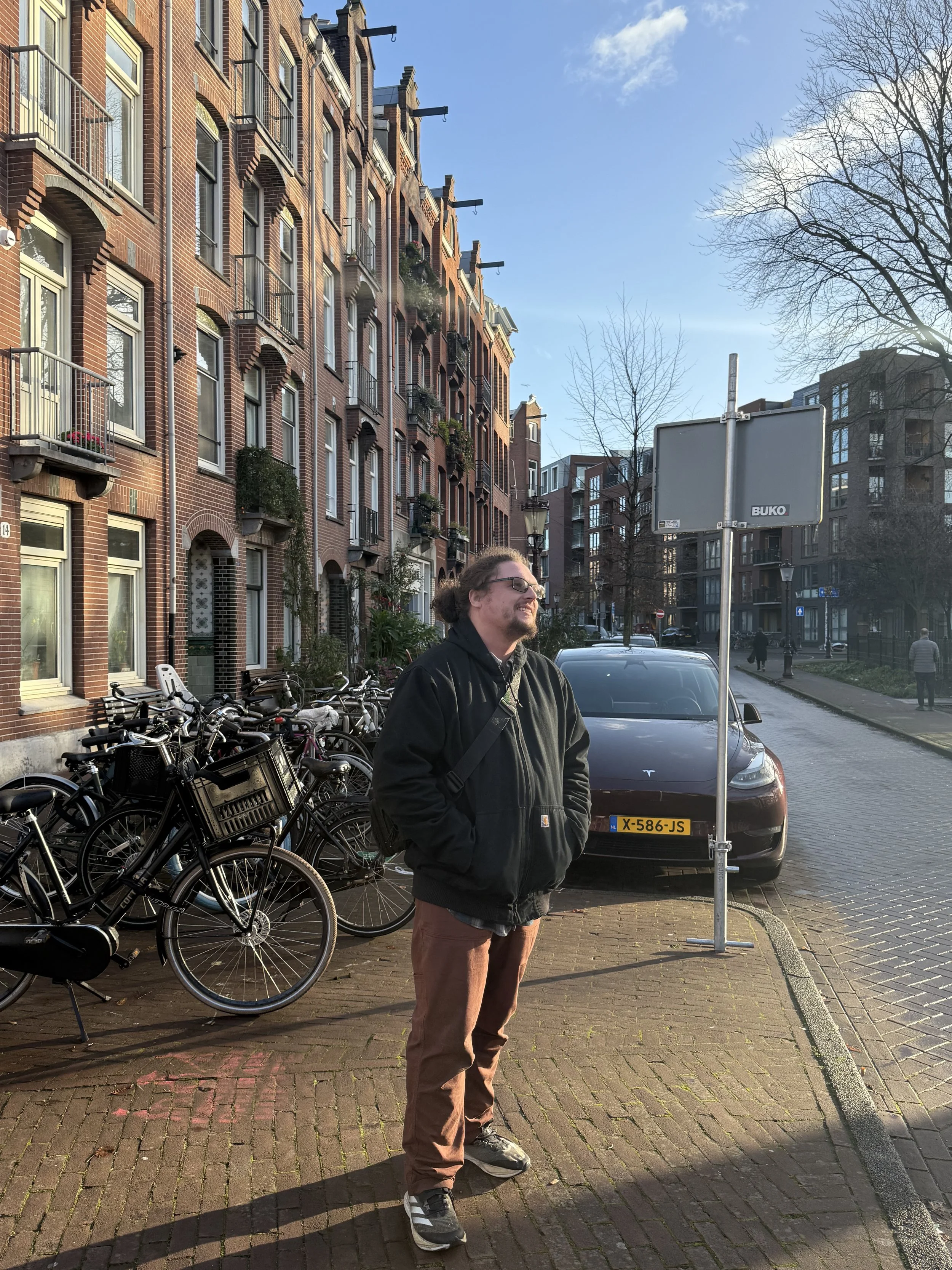 A man wearing glasses, a black jacket, and brown pants, with a backpack, is standing on a brick sidewalk, smiling with eyes closed, in front of a row of bicycles and a dark-colored Tesla car, in an urban street with brick buildings and leafless trees