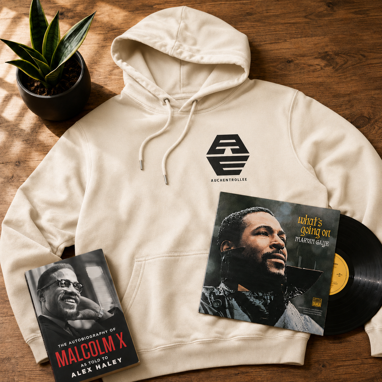 A beige hoodie with the logo and text 'AUCHENTROLLEE' on it laid out on a wooden surface. To the right of the hoodie, there is a vinyl record and a black book titled 'The Autobiography of Malcolm X as Told to Alex Haley.' To the right of the hoodie, there is a vinyl album with a black and white cover showing a man wearing glasses, and the title 'What's Going On' by Marvin Gaye. There is also a potted snake plant on the upper left corner.