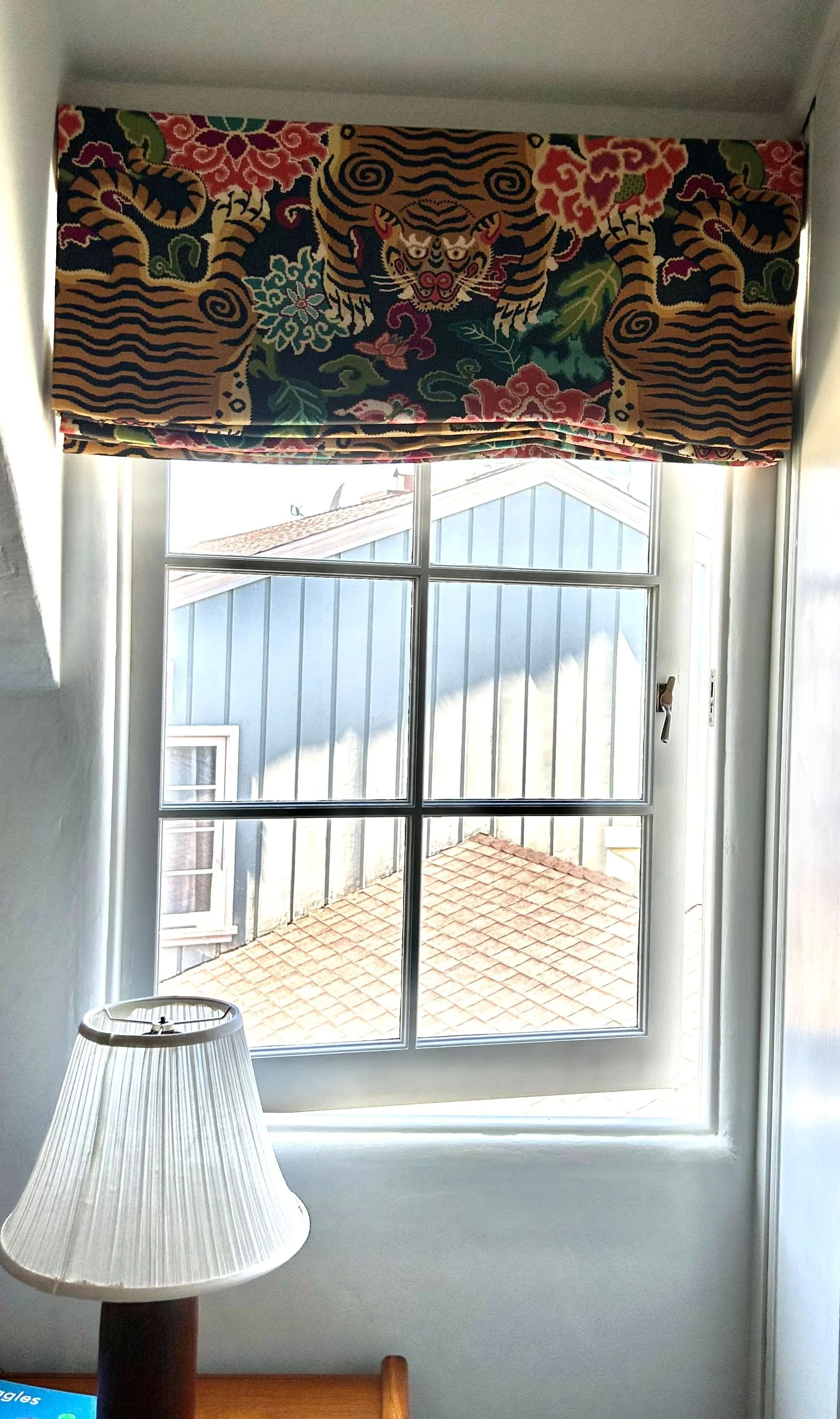A window with a decorative curtain featuring a tiger and floral pattern, a table lamp, and a portion of a wooden furniture piece.