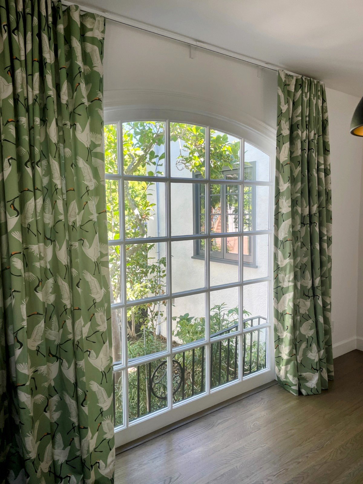A large window with white grid panes and rounded top, flanked by green curtains with a white crane pattern, looking out onto a garden with trees and plants.