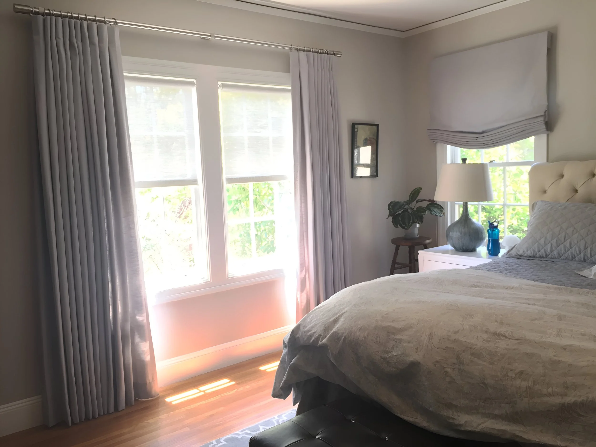 A bedroom with a bed, nightstand, lamp, houseplant, windows, and curtains.