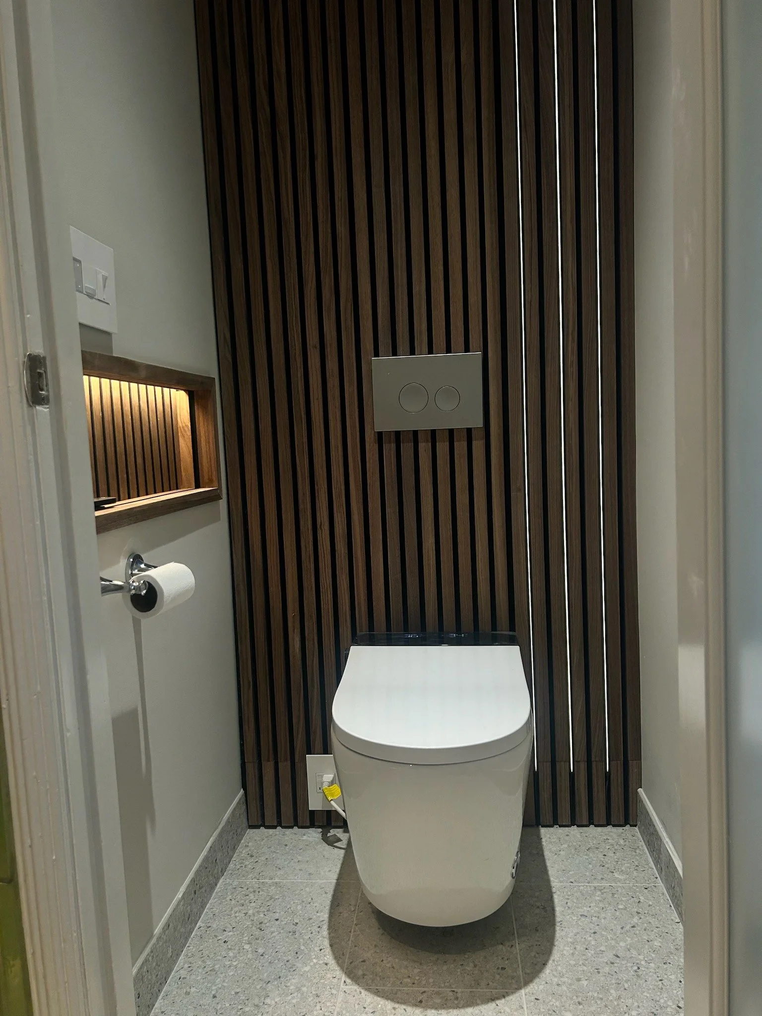 Modern bathroom with a smart toilet, black and brown wooden vertical acoustic panels, lighted toilet paper holder niche.