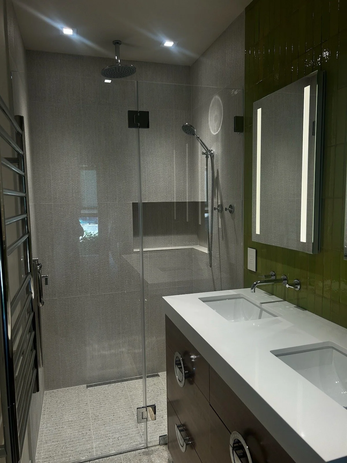 Modern bathroom with walk-in shower featuring a rain showerhead and handheld shower, a white double sink vanity with a dark wood cabinet, a green tiled wall with a large mirror, and a heated towel rail.