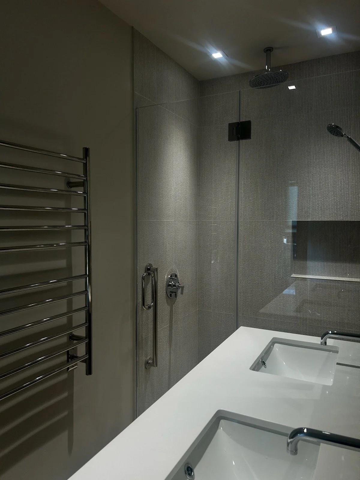 Modern bathroom featuring a glass-enclosed shower with a rain shower head, a white sink countertop with integrated sinks, a wall-mounted faucet, and a stainless steel heated towel rack.