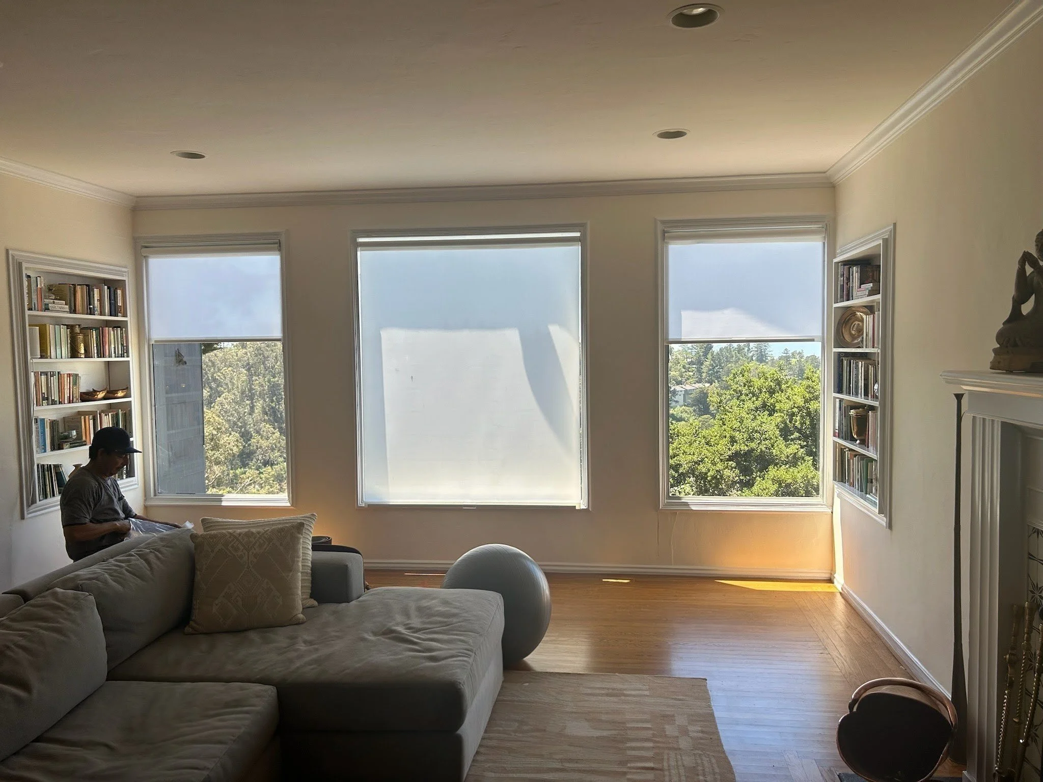 A bright living room with three large windows overlooking trees. There's a sofa with cushions, a round object on the floor, bookshelves on both sides of the windows, and a person sitting on the left side reading.