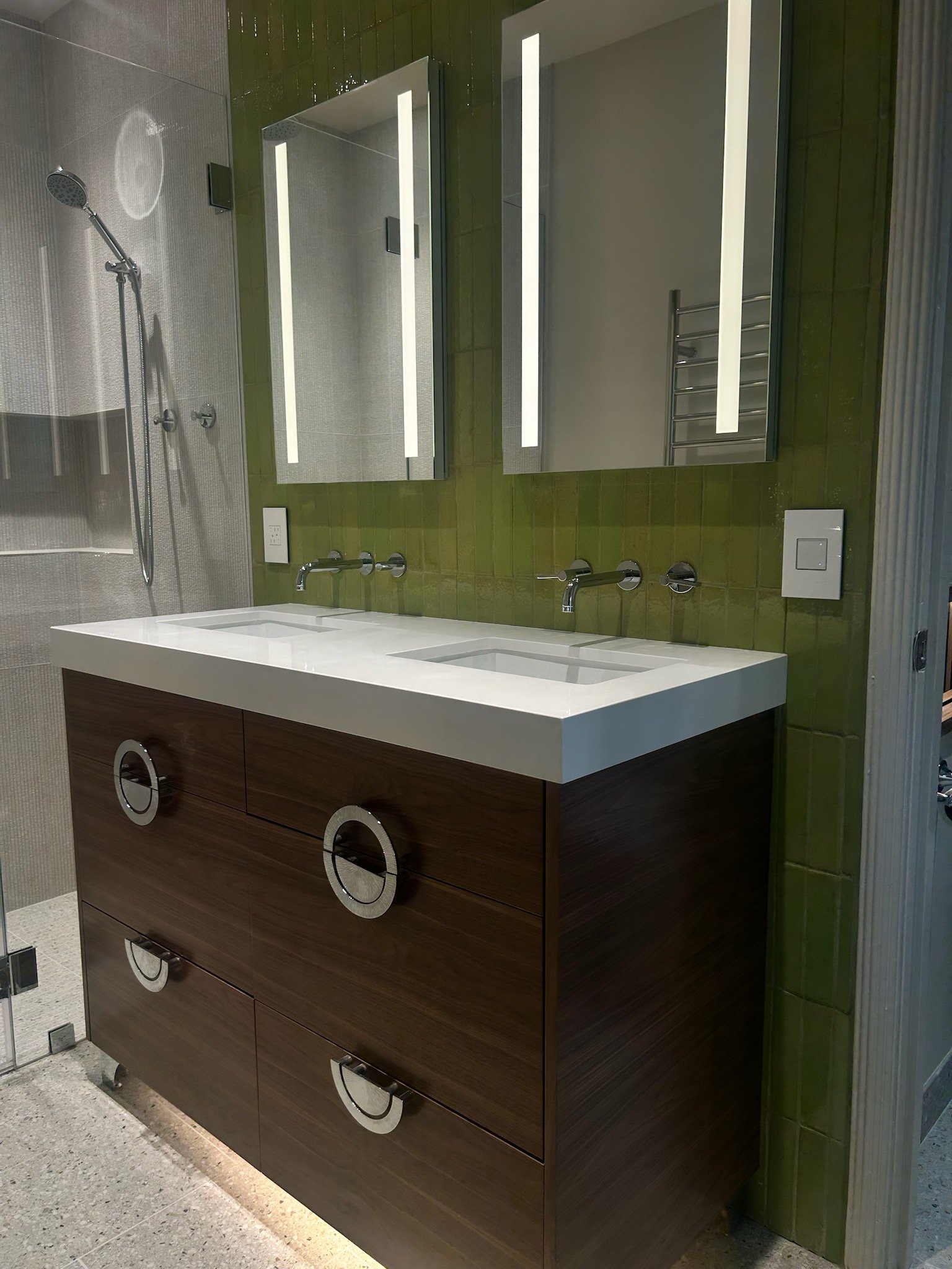 Modern bathroom with green tiled wall, large mirror with LED lighting, custom double sink vanity in wood finish, and a shower with a rainhead showerhead.