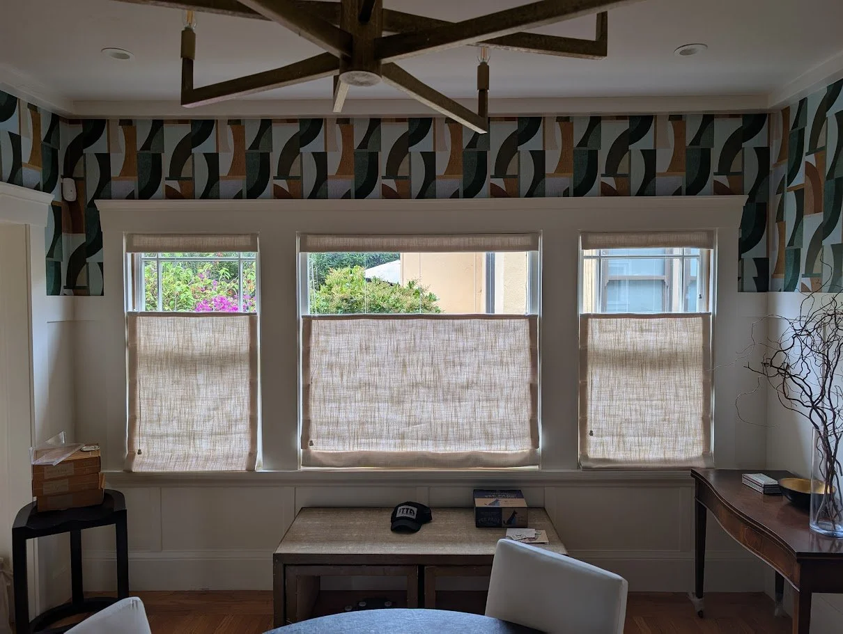 Room with three windows covered by tan blinds, a decorative wallpaper border with geometric pattern, a wooden ceiling fan, a small table with a box and cap, a desk with papers, and a vase with twigs.