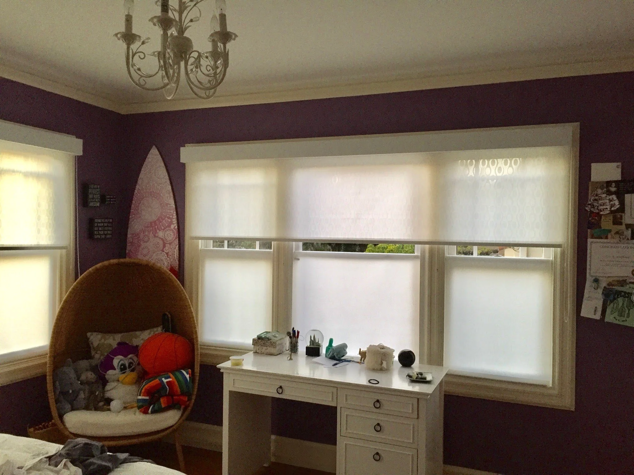 A bedroom with purple walls, three large windows with white blinds, a wicker chair with stuffed animals and a blanket, and a white desk with miscellaneous items on it. Chandelier overhead.