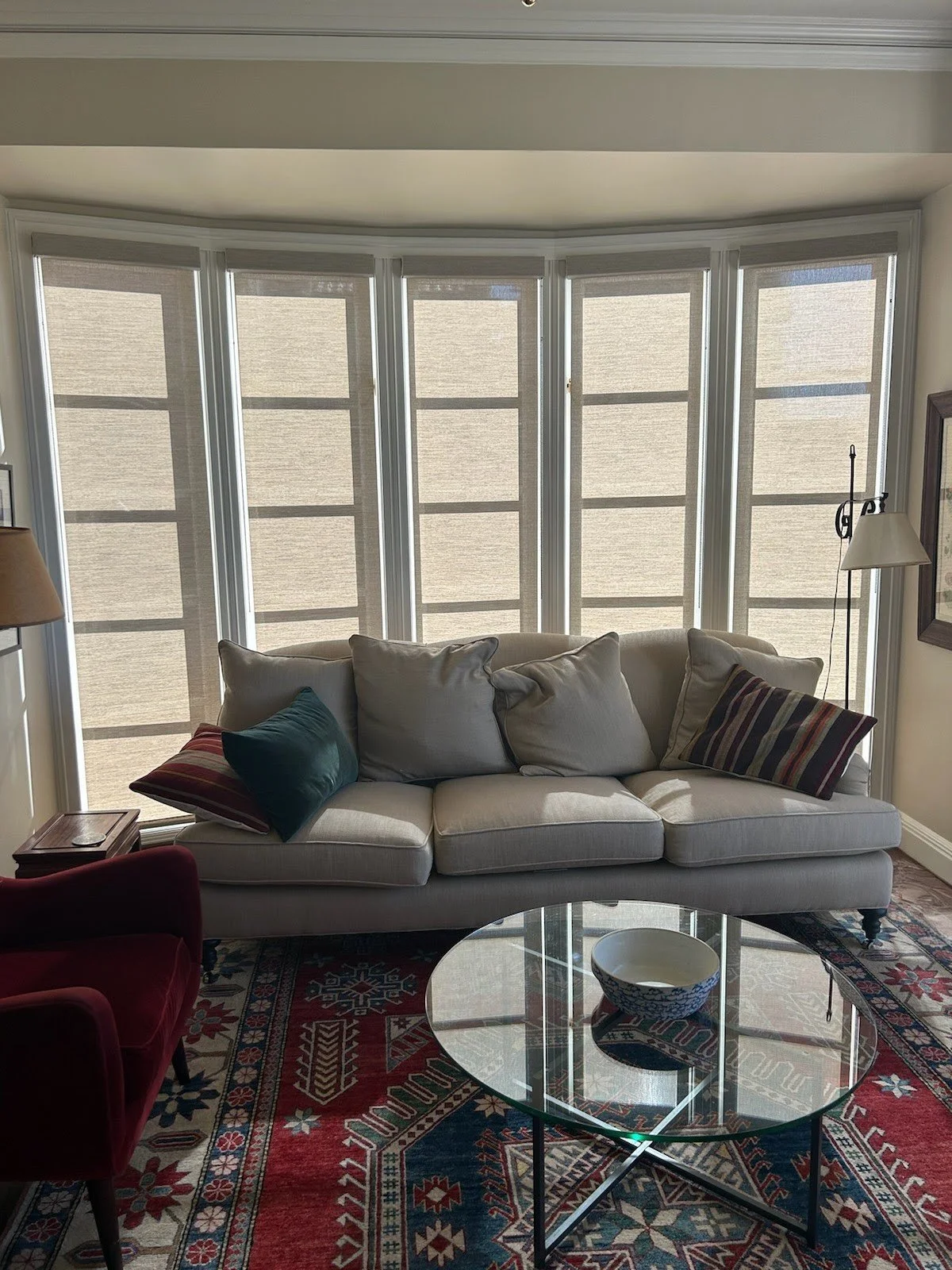 Living room with beige sofa, colorful pillows, glass coffee table with a bowl, red armchair, and bay windows with beige shades.