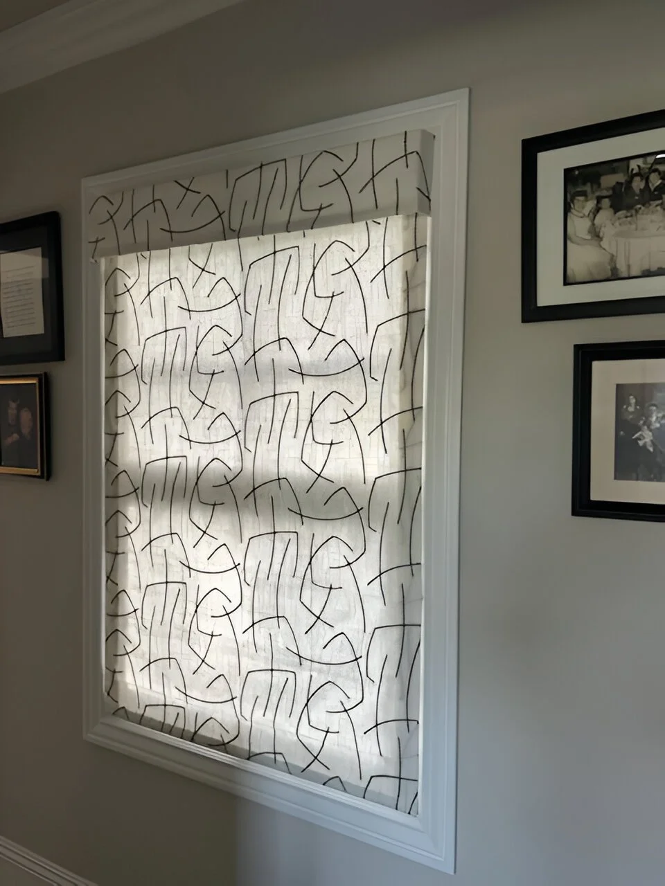 Window with patterned curtain featuring abstract black lines on a white background, surrounded by framed photographs on a white wall.