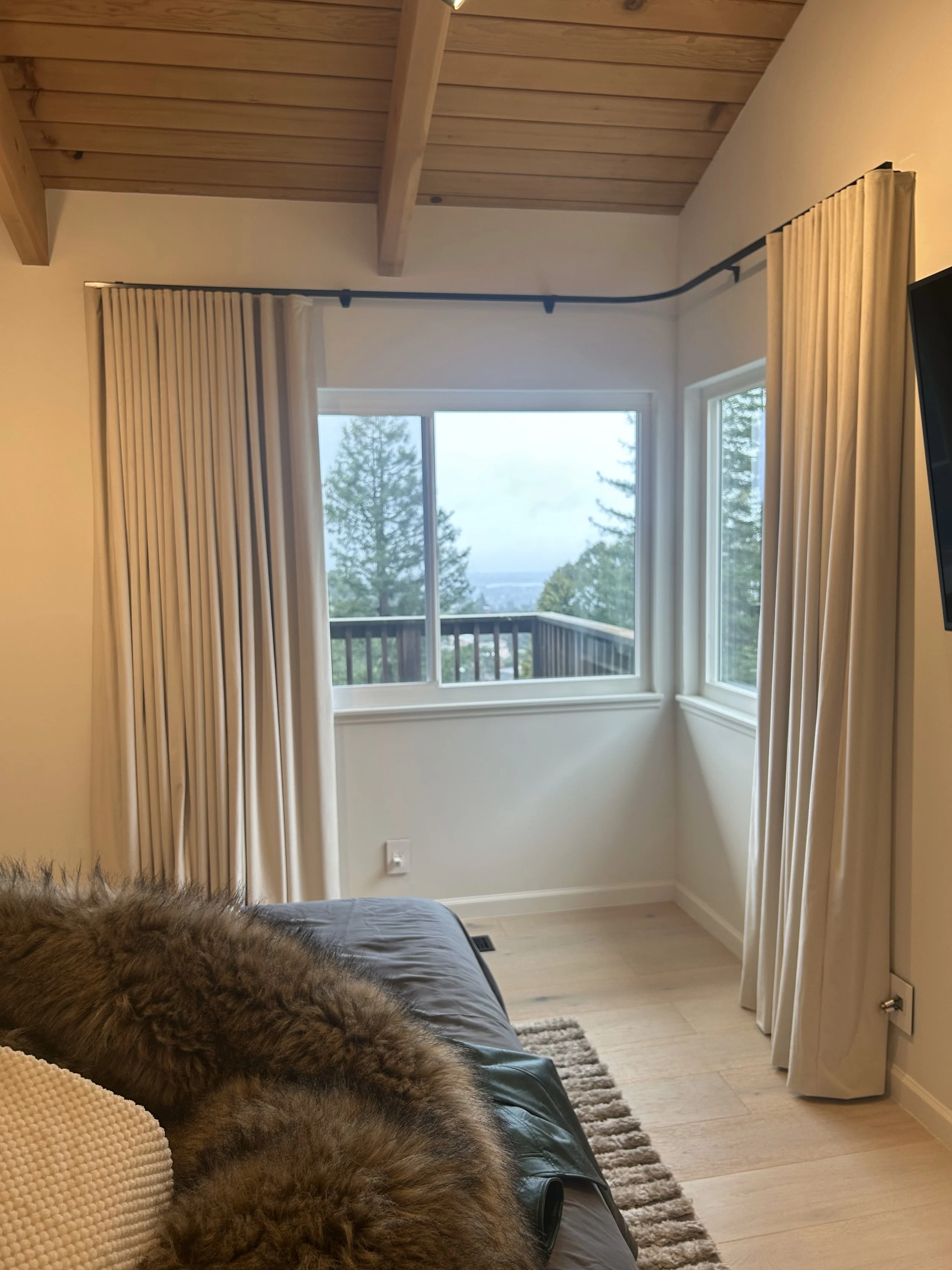 Living room with large windows showing trees and a balcony, beige curtains, a dark brown leather couch with a faux fur blanket, a beige pillow, and a corner of a wall-mounted TV.