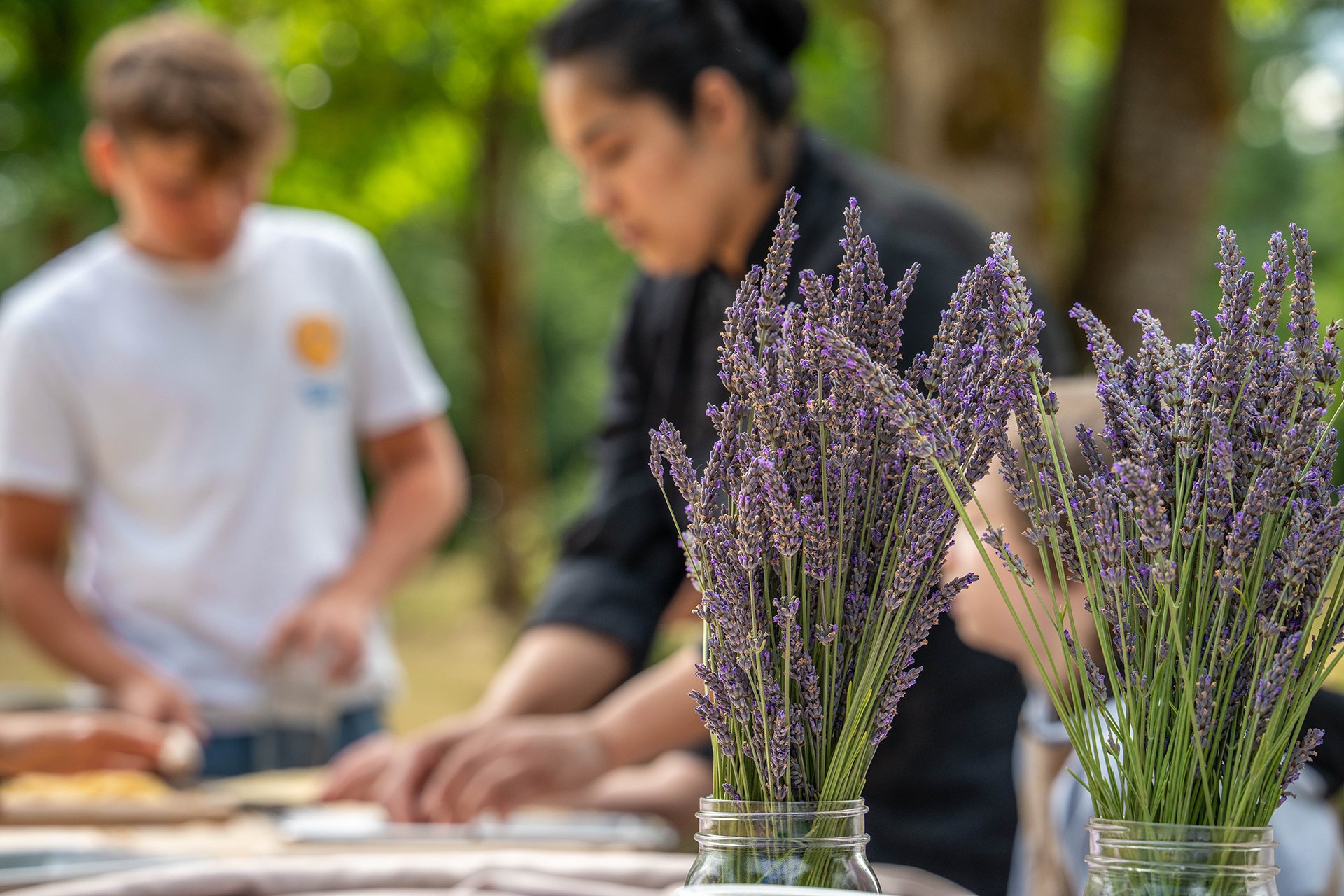Two jars of fresh lavender on a table with a woman teaching children out of focus in the background.