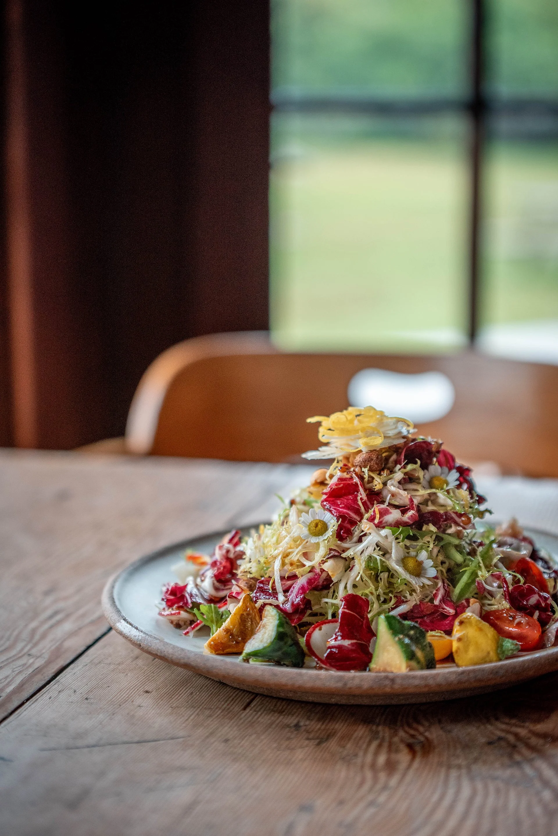 A towering salad with edible flowers sits on a wooden table in front of a window.