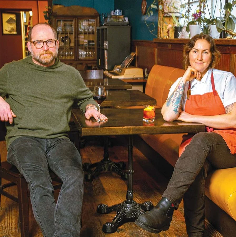 A man and woman sit at a table showcasing a glass of wine and cocktail.