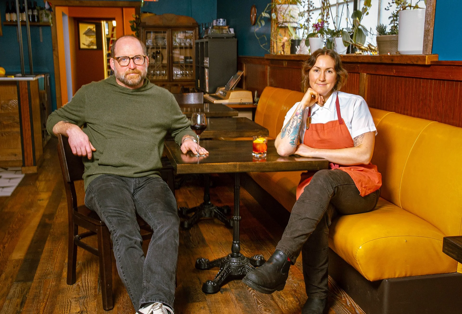 A man and woman, a chef, sitting at a table in a restaurant with a glass of wine and a cocktail in front of them.