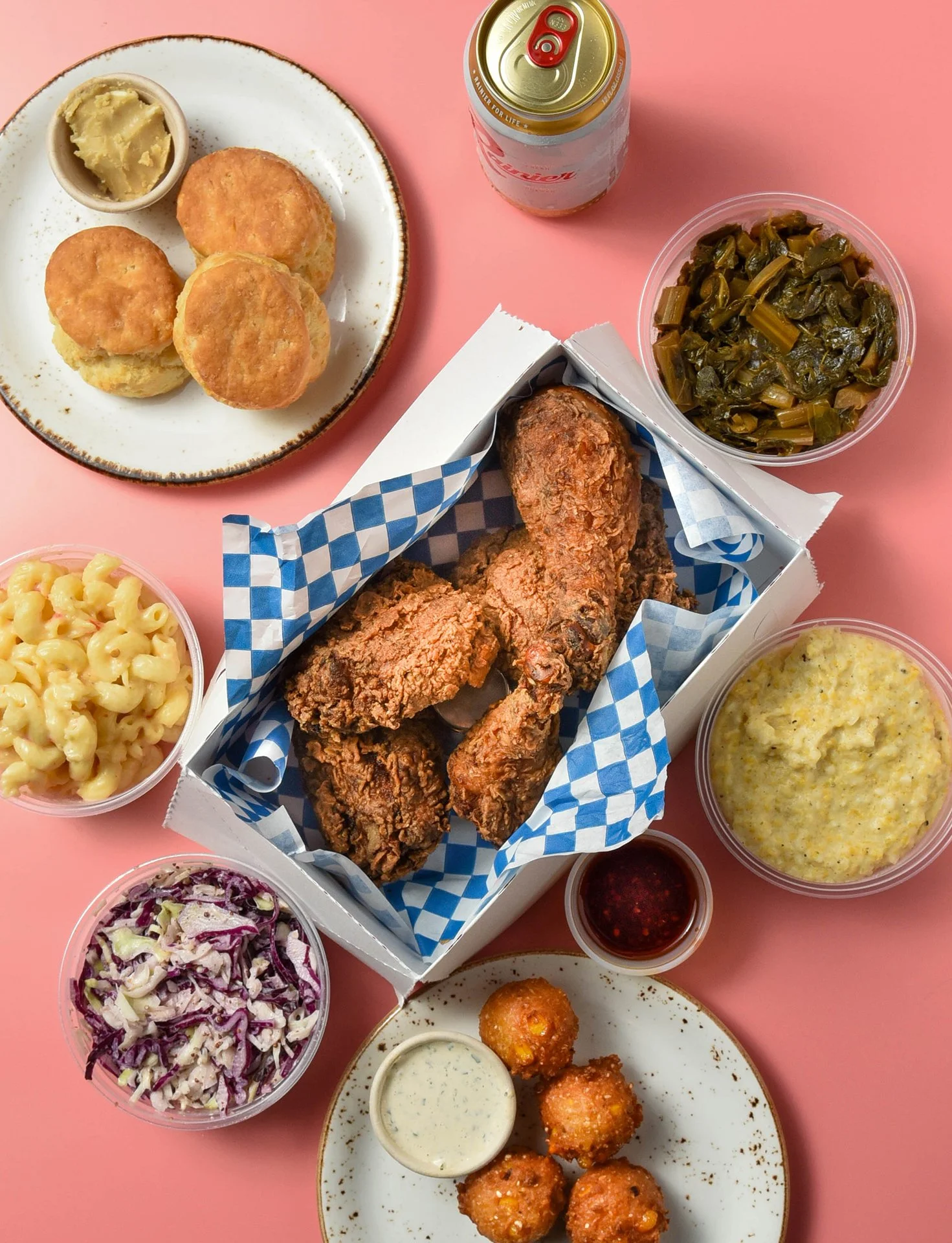 An top down image of a fried chicken dinner featuring biscuits, mac and cheese, coleslaw, hush puppies, grits, greens, sauces, and a can of beer