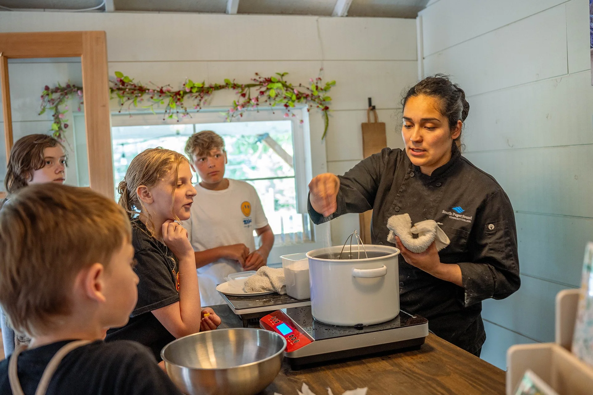 A woman sprinkles salt into a pot of boiling water as children look on.