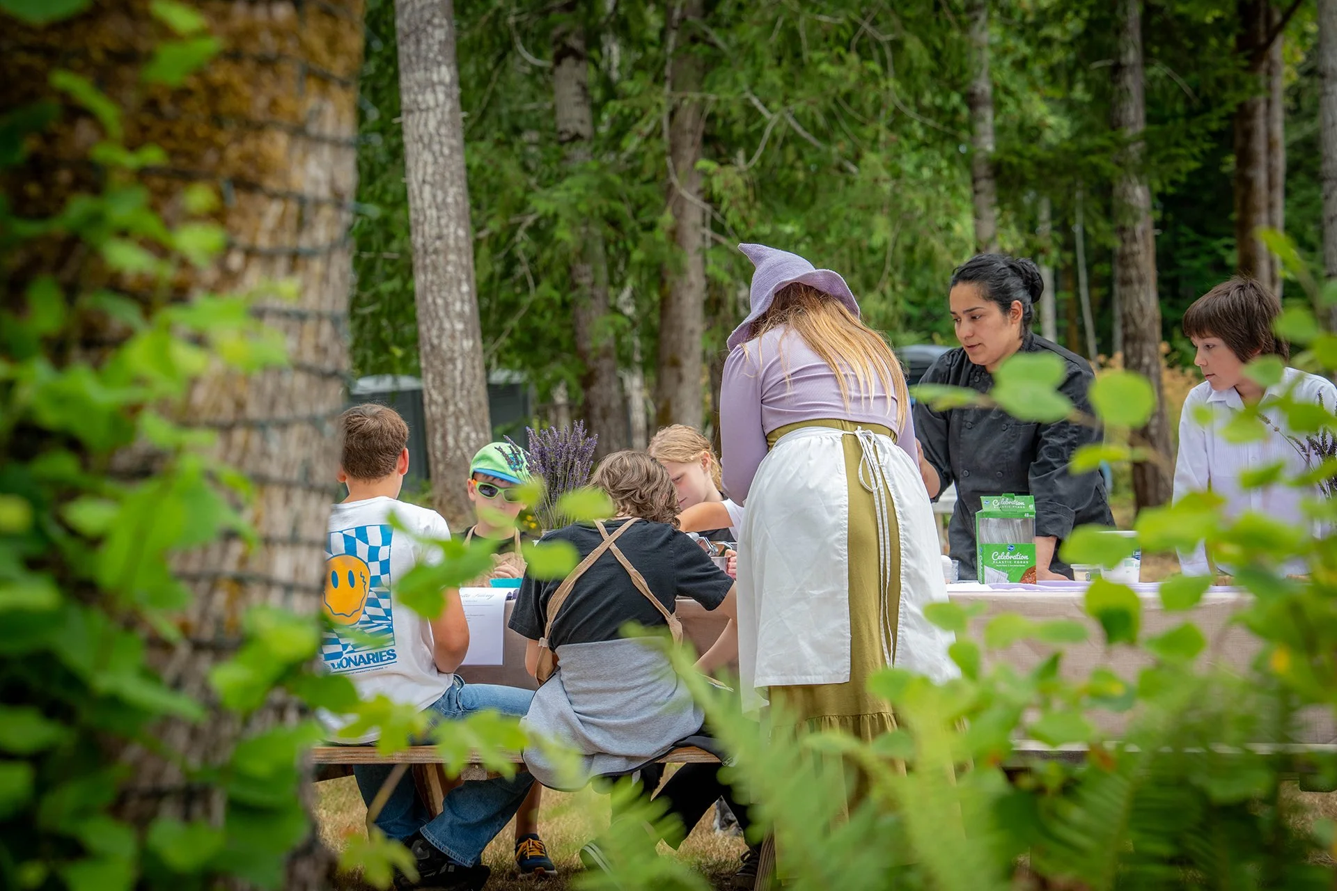 Children sitting at a table outdoors surrounded by green trees.