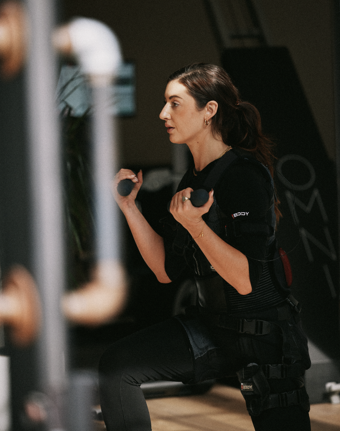 A woman in black workout clothes is doing squats with dumbbells in a gym.