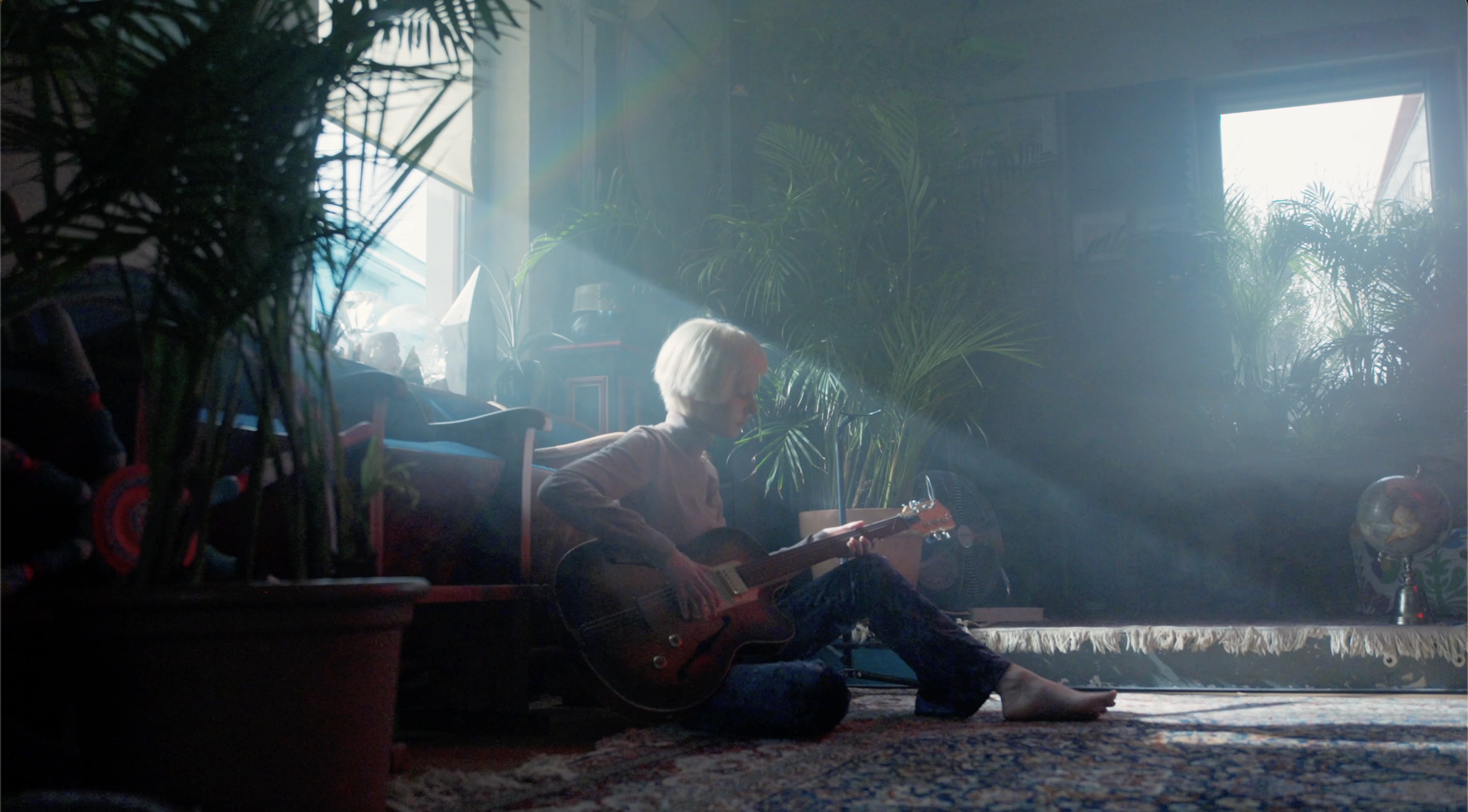A person with short white hair, sitting on the floor, playing an acoustic guitar, surrounded by lush green plants, with sunlight streaming through a window in a cozy interior setting.