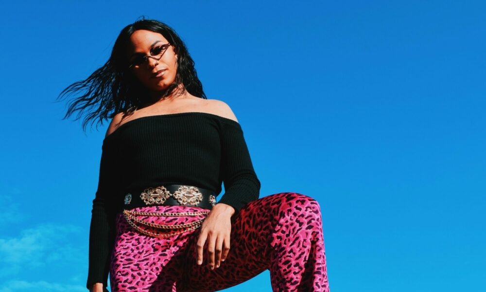 A person wearing sunglasses, a black off-the-shoulder top, and vibrant pink leopard-print pants with a decorative belt, standing outdoors against a bright blue sky.