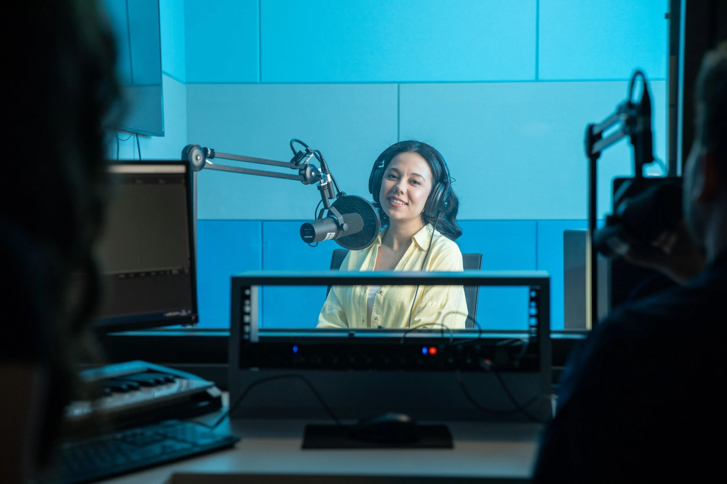 A woman wearing headphones sitting in front of a microphone in a recording studio, with a bright blue wall behind her. There are people and equipment partially visible in the foreground and on the sides.