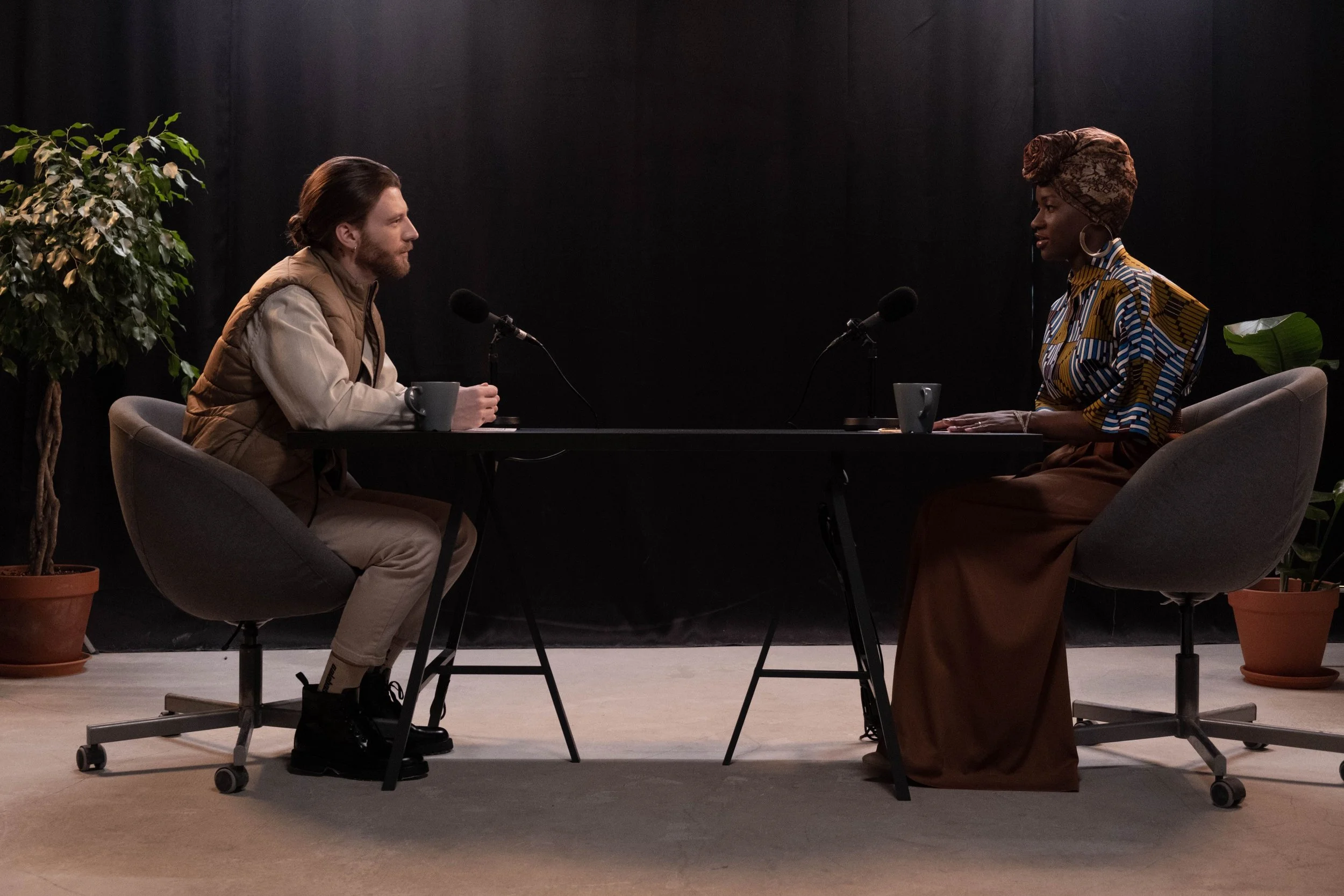 A man and woman sit across from each other at a table with microphones, engaging in a conversation or interview; there are two potted plants in the background.
