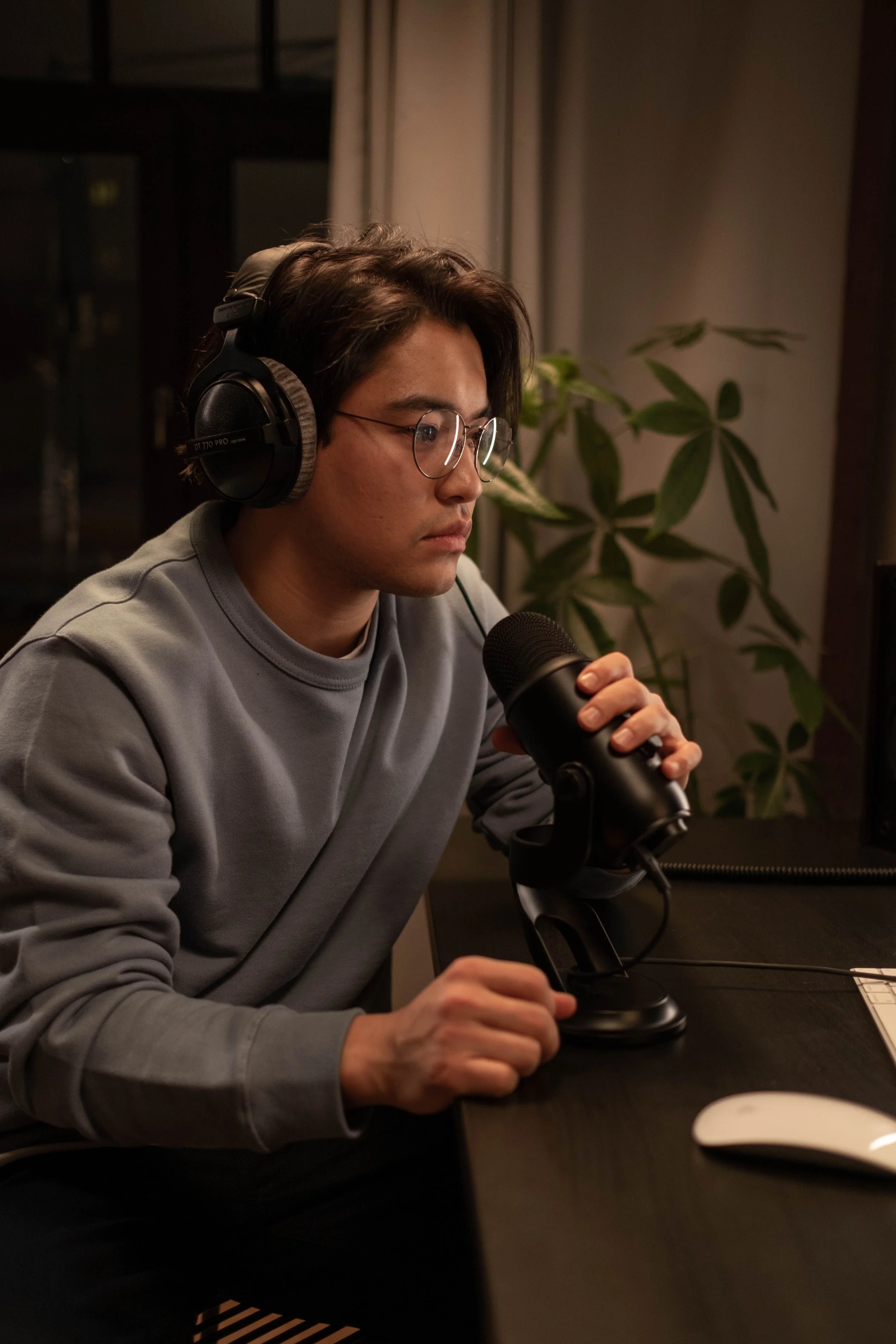 A young man with glasses and dark hair, wearing a gray sweatshirt, is sitting at a desk in front of a microphone. He is wearing over-ear headphones and holding the microphone with one hand. There is a potted plant and a white curtain in the background, with dim lighting.