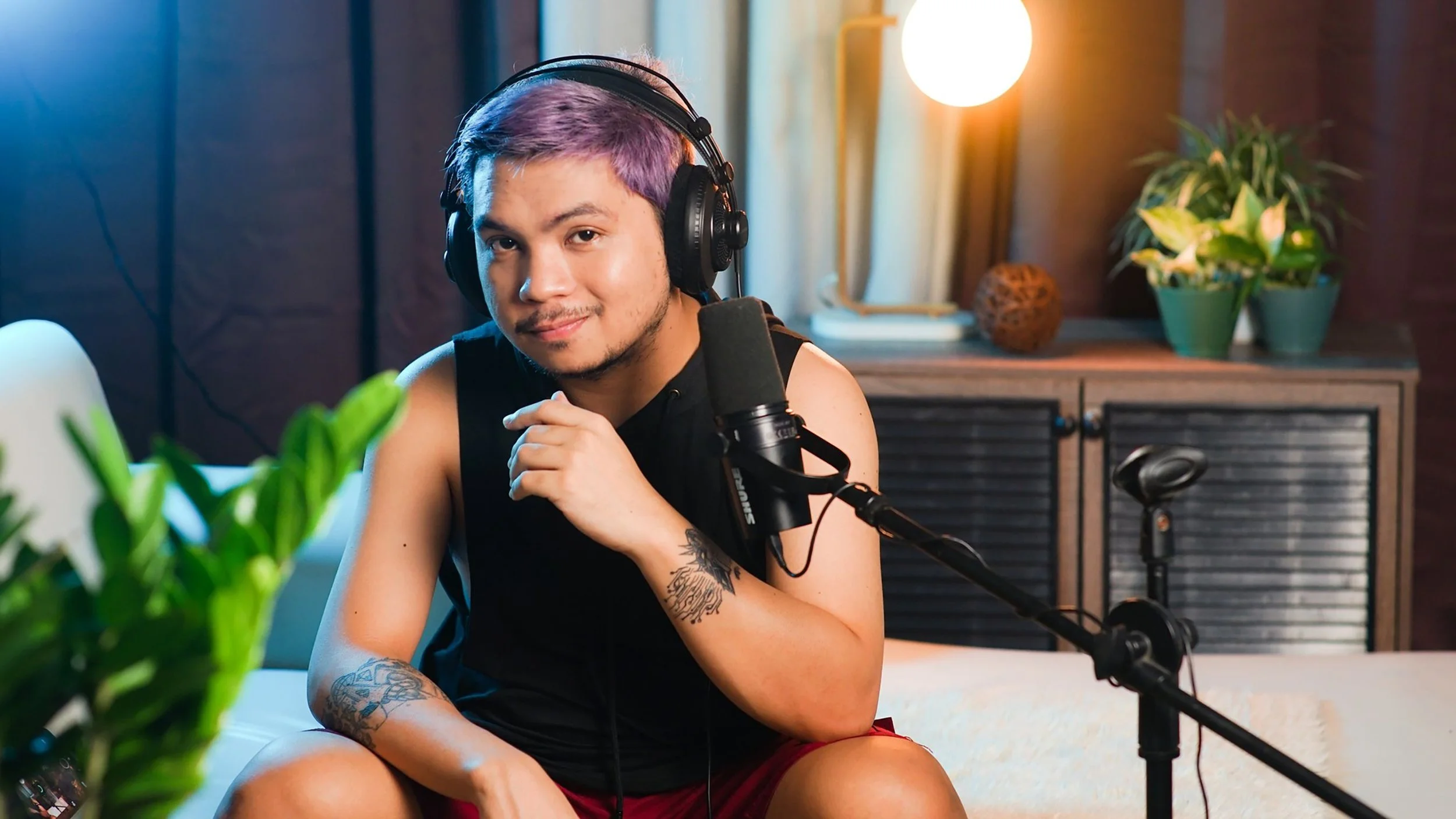 A person with purple hair, wearing headphones, sitting at a desk with a microphone, smiling at the camera in a cozy room with plants and warm lighting.