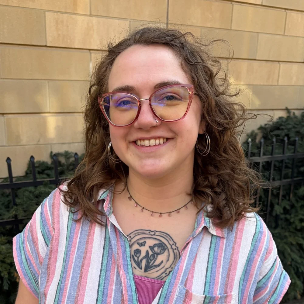 A young woman with curly hair, glasses, and a nose piercing smiling outdoors in front of a brick wall and black metal fence.