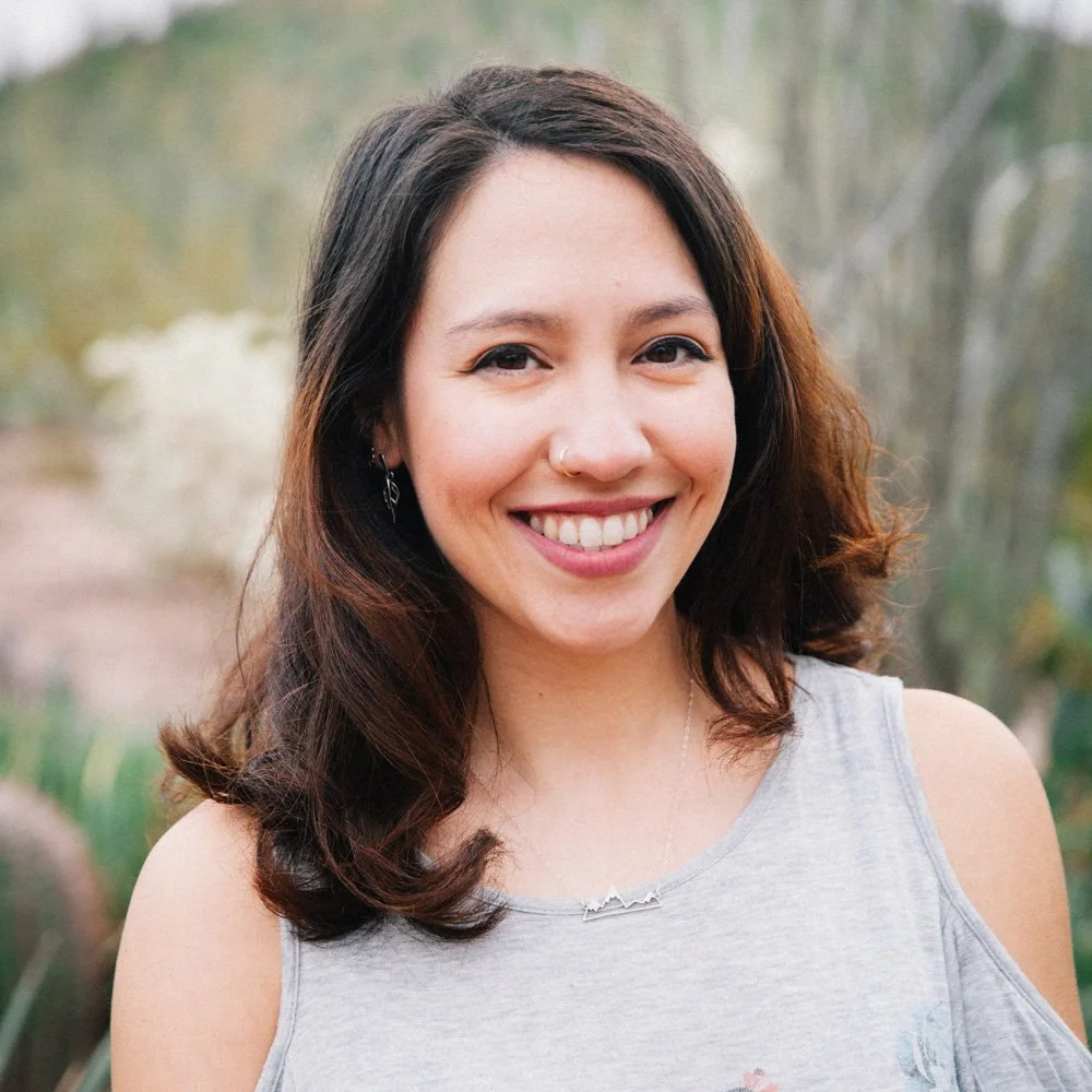 A young woman with shoulder-length brown hair smiling outdoors, wearing a gray off-shoulder top and jewelry, with a blurred natural background.