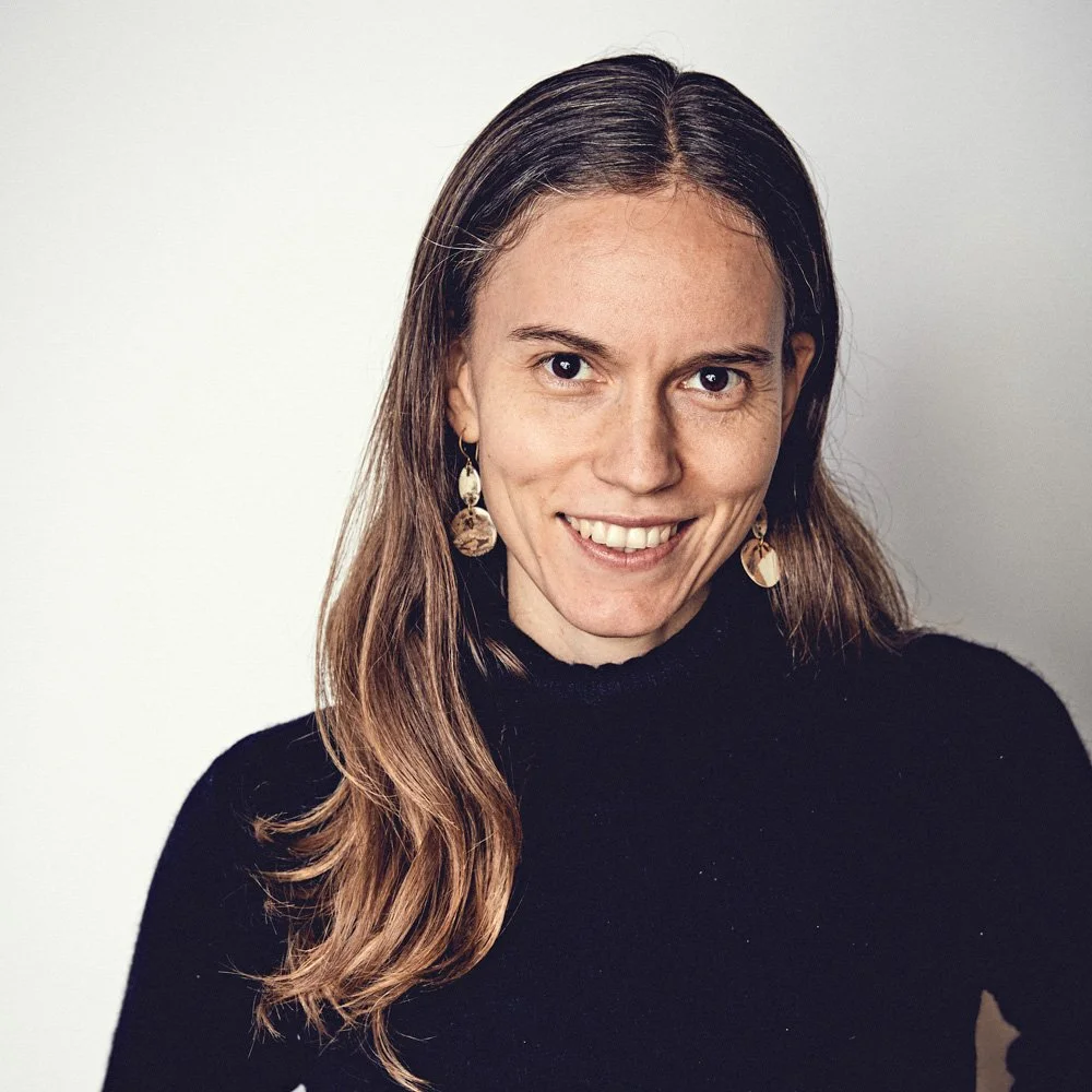 A woman with long brown hair and earrings smiling at the camera against a plain background.