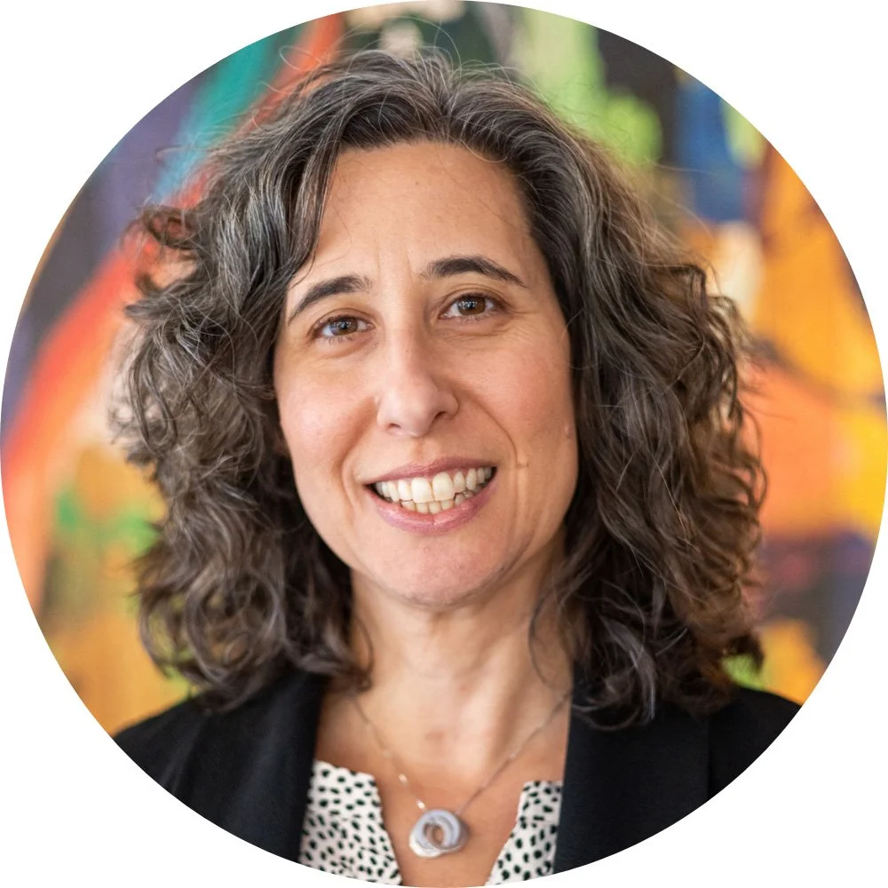 Emily Weisgrau smiling at the camera, wearing a black blazer and necklace with curly shoulder-length hair.