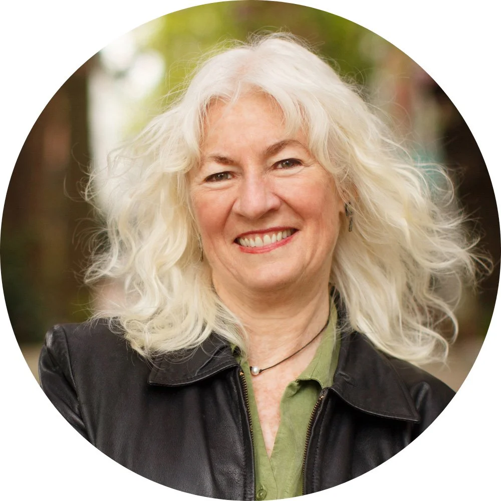 Judy Wicks smiling with long, curly white hair, wearing a black leather jacket and a pendant necklace.