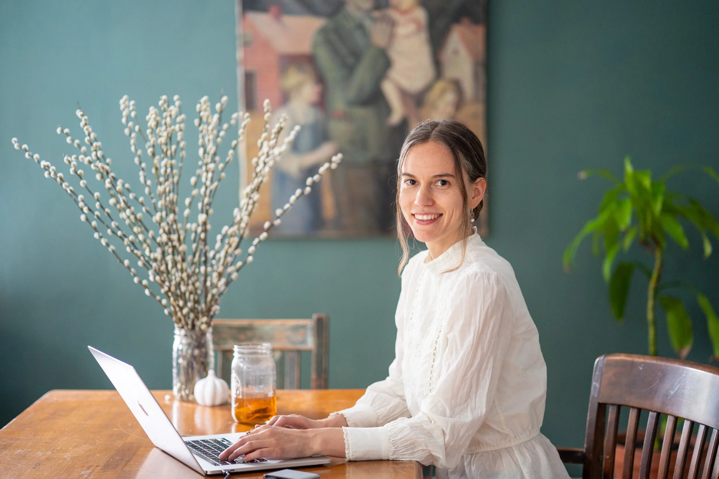 A woman with long brown hair, wearing a white blouse, sitting at a wooden table working on a silver laptop, with a large vase of pussy willow branches, a small white pumpkin, and a glass of amber-colored drink in front of her, in a room with teal walls and a painting of children playing in the background.