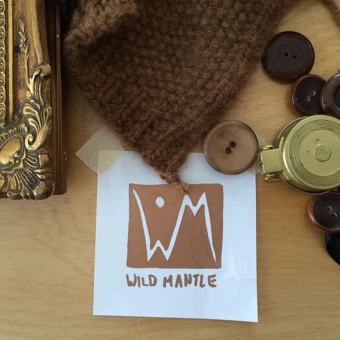 A brown knitted hat, several wooden buttons, a green metal container, and a printed logo with the words "Wild Mante" on a wooden surface.