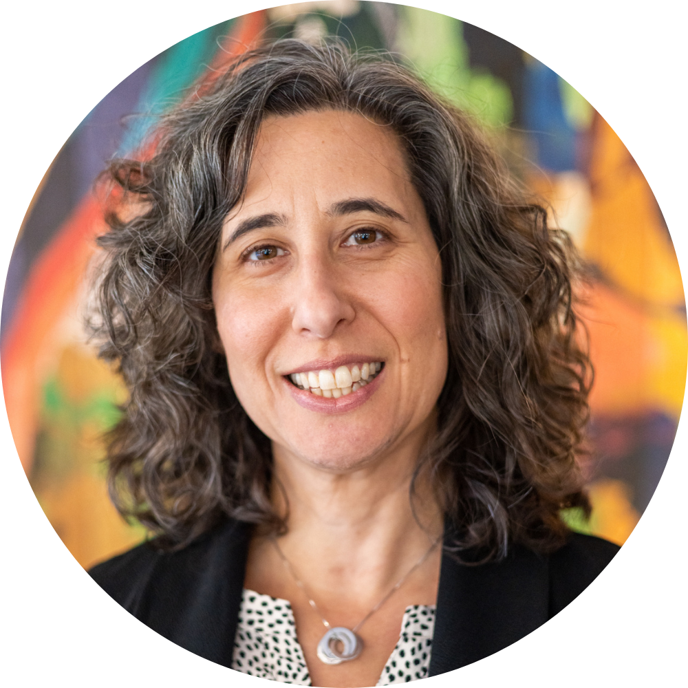 Emily Weisgrau smiling at the camera, wearing a black blazer and necklace with curly shoulder-length hair.