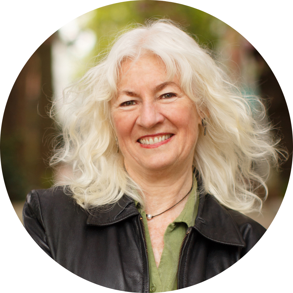 Judy Wicks smiling with long, curly white hair, wearing a black leather jacket and a pendant necklace.