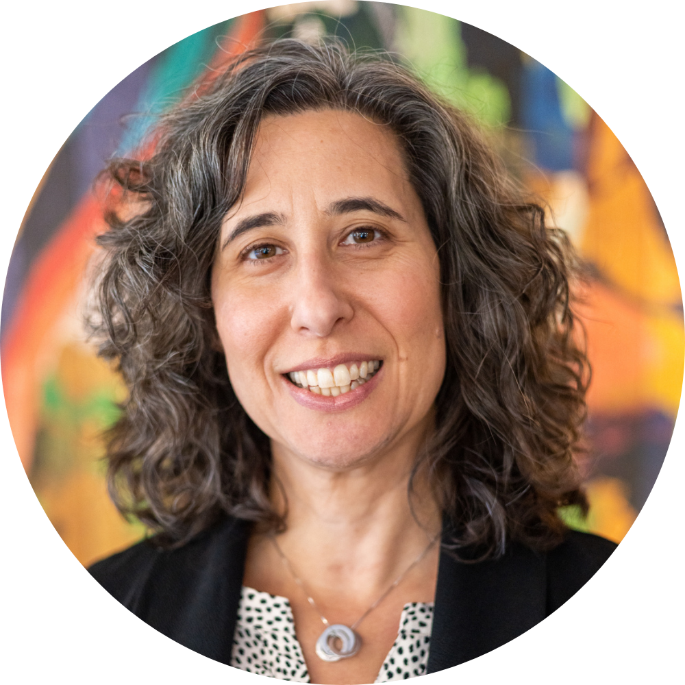 Emily Weisgrau smiling at the camera, wearing a black blazer and necklace with curly shoulder-length hair.