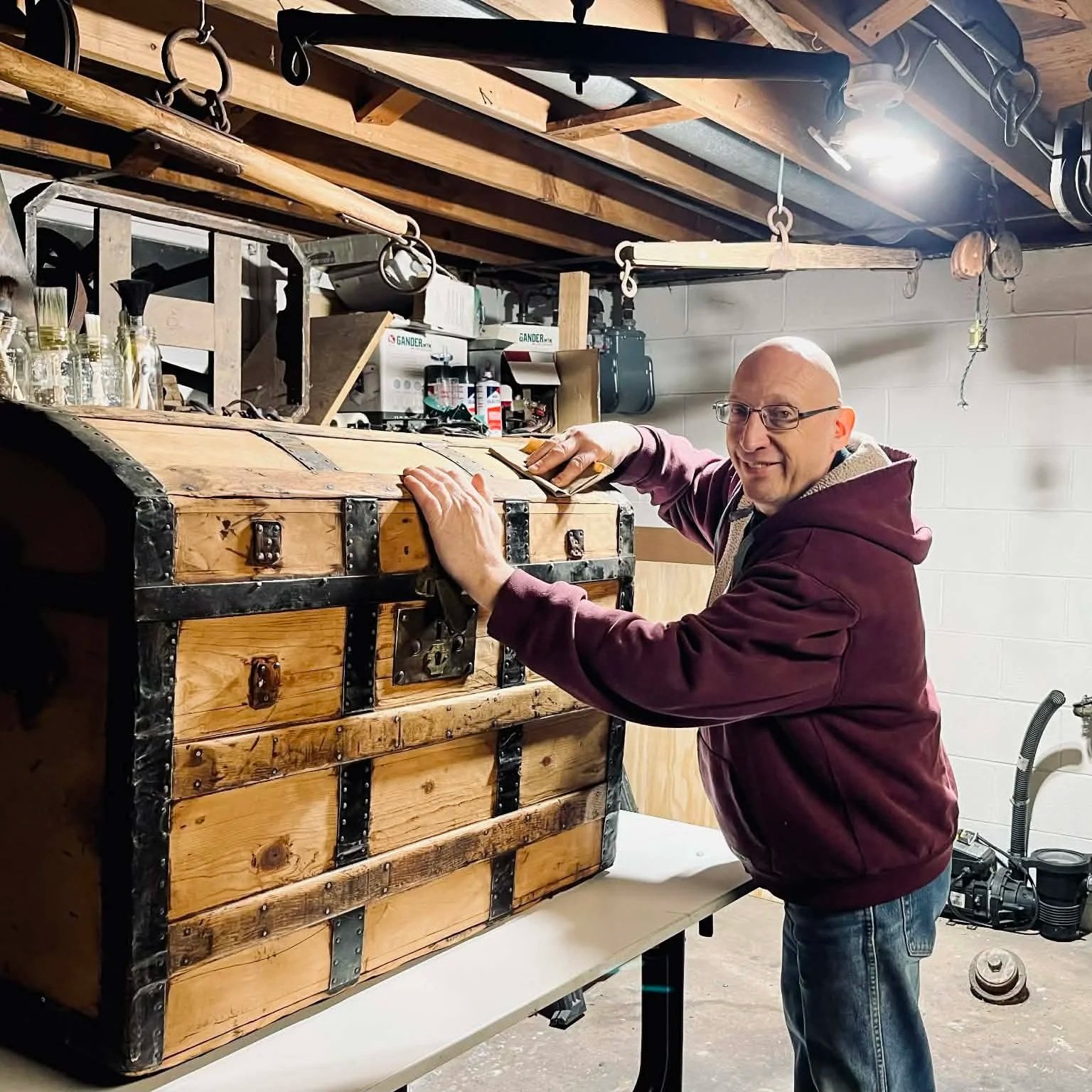 A man in a maroon hoodie and glasses is polishing an antique wooden chest with black metal accents in a workshop.