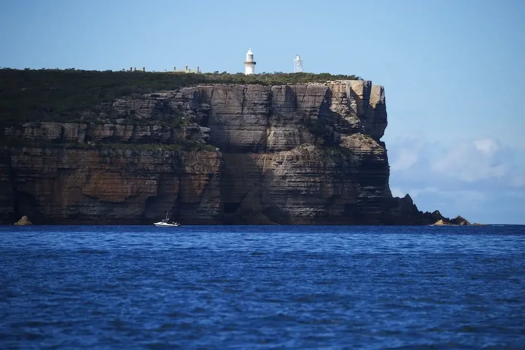 A large ocean cliff with a lighthouse and antenna on top, with a sailboat on the water below.