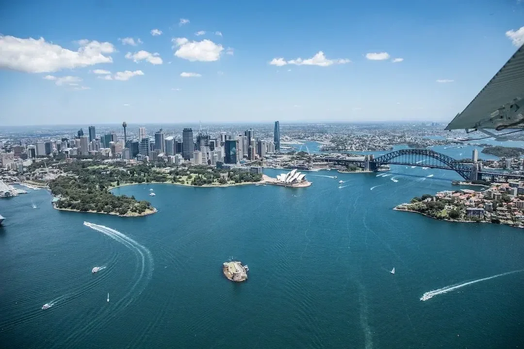 Aerial view of Sydney skyline featuring tall modern skyscrapers, the Sydney Opera House, and Harbour Bridge with boats sailing on the water under a partly cloudy blue sky.