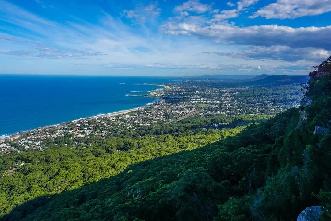 A scenic view from a mountain overlooking a coastal town with green forests, a beach, and a harbor under a partly cloudy sky.