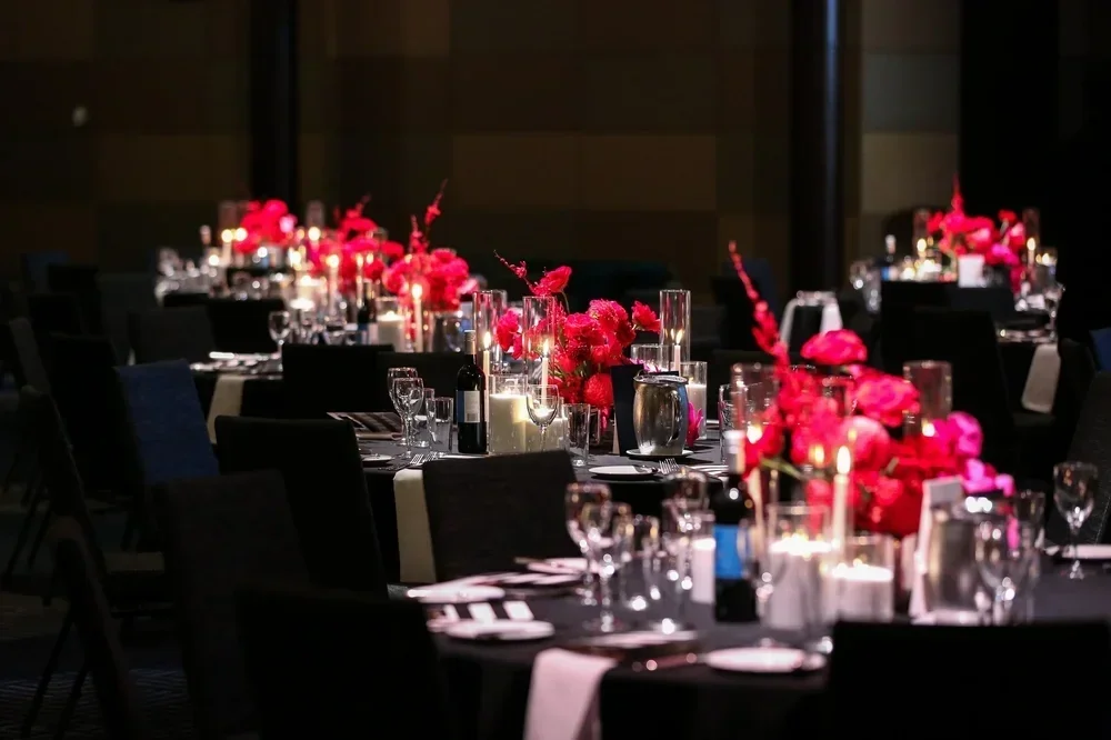 Elegant event table setup with black chairs, floral centerpieces with red flowers, candles, and tableware in a dimly lit room.